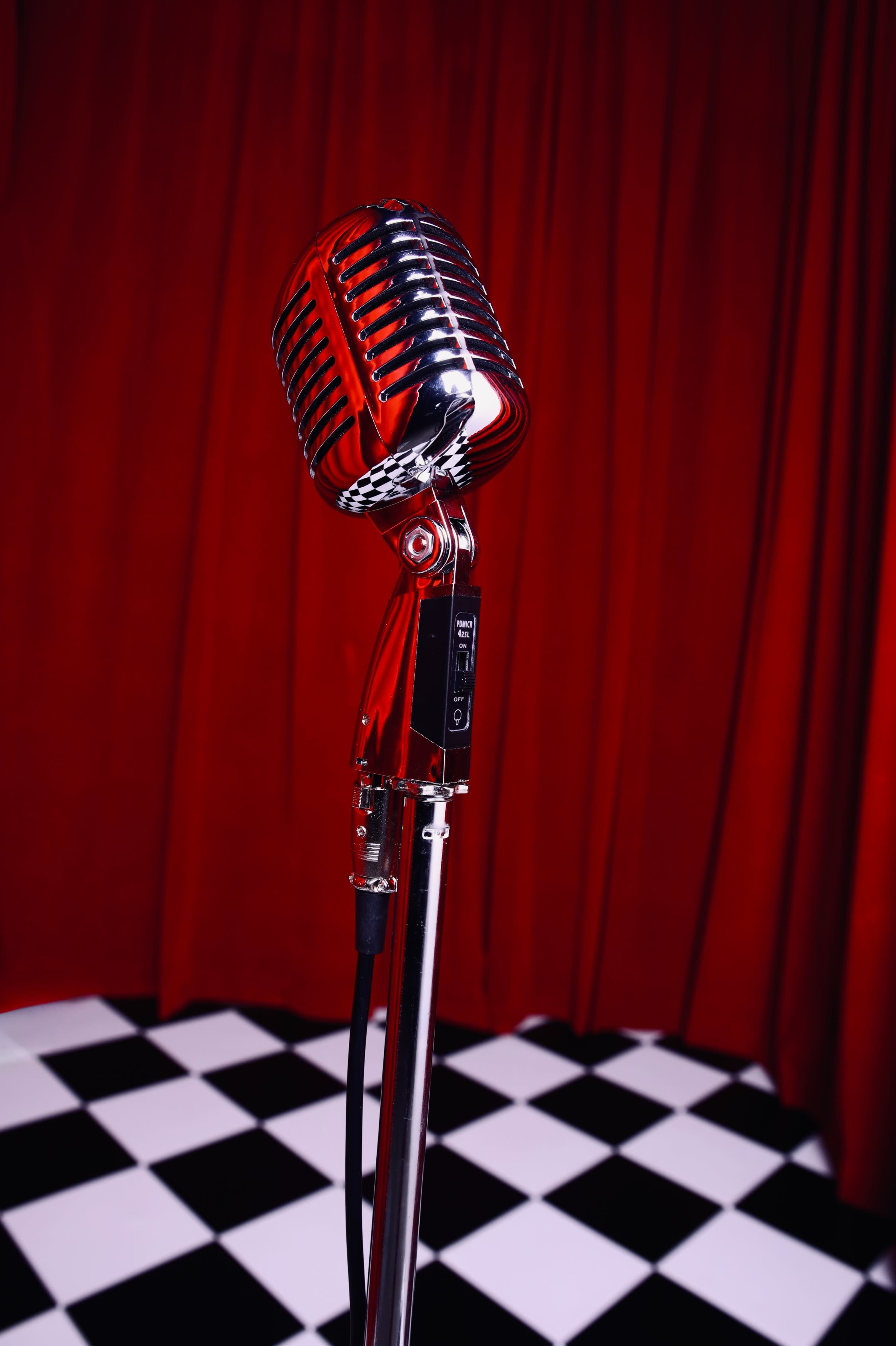 A vintage-style microphone stands on a black and white checkered floor in front of a red curtain.