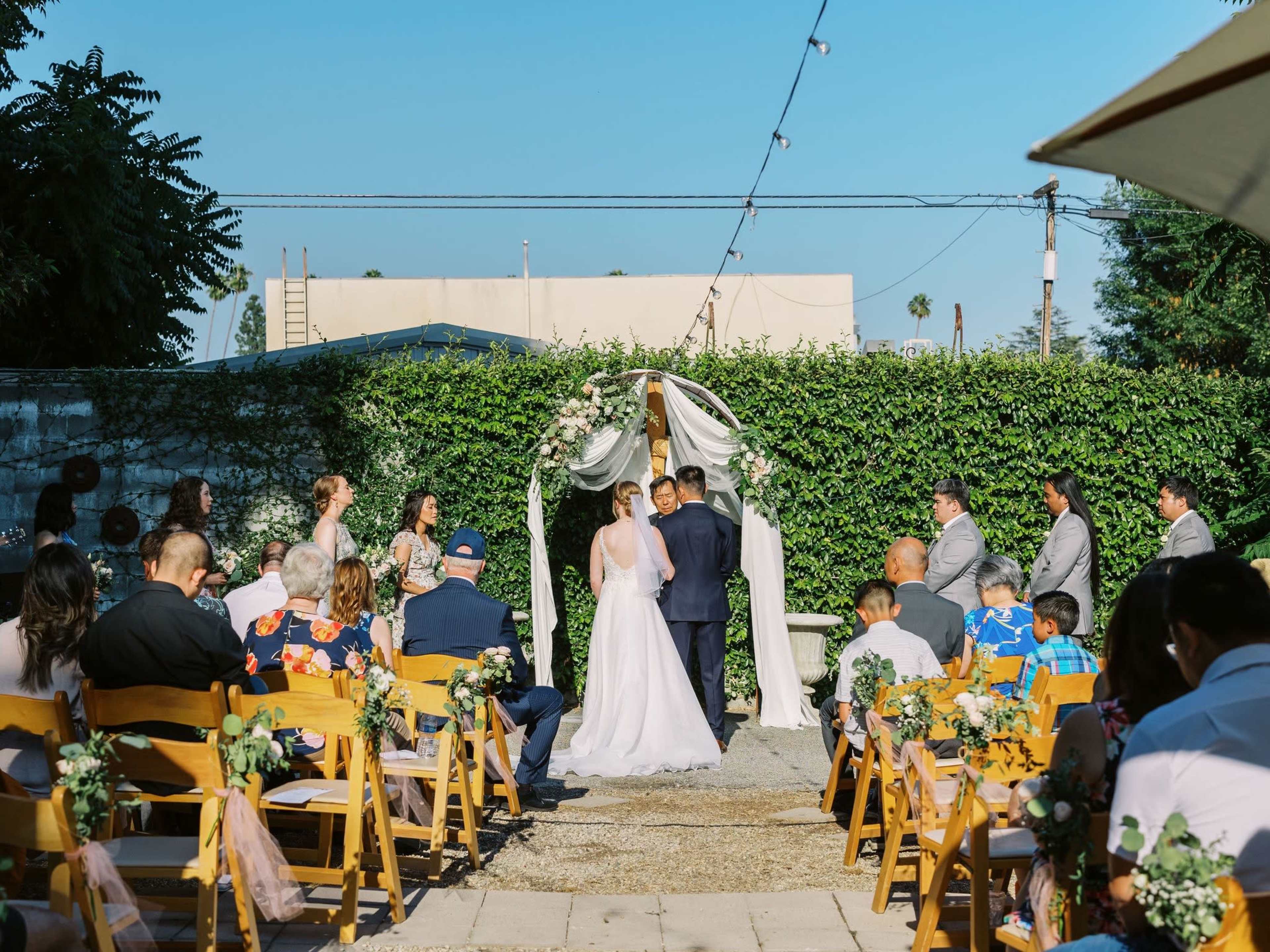 A couple stands under a decorated arch exchanging vows in front of seated guests at an outdoor wedding ceremony.