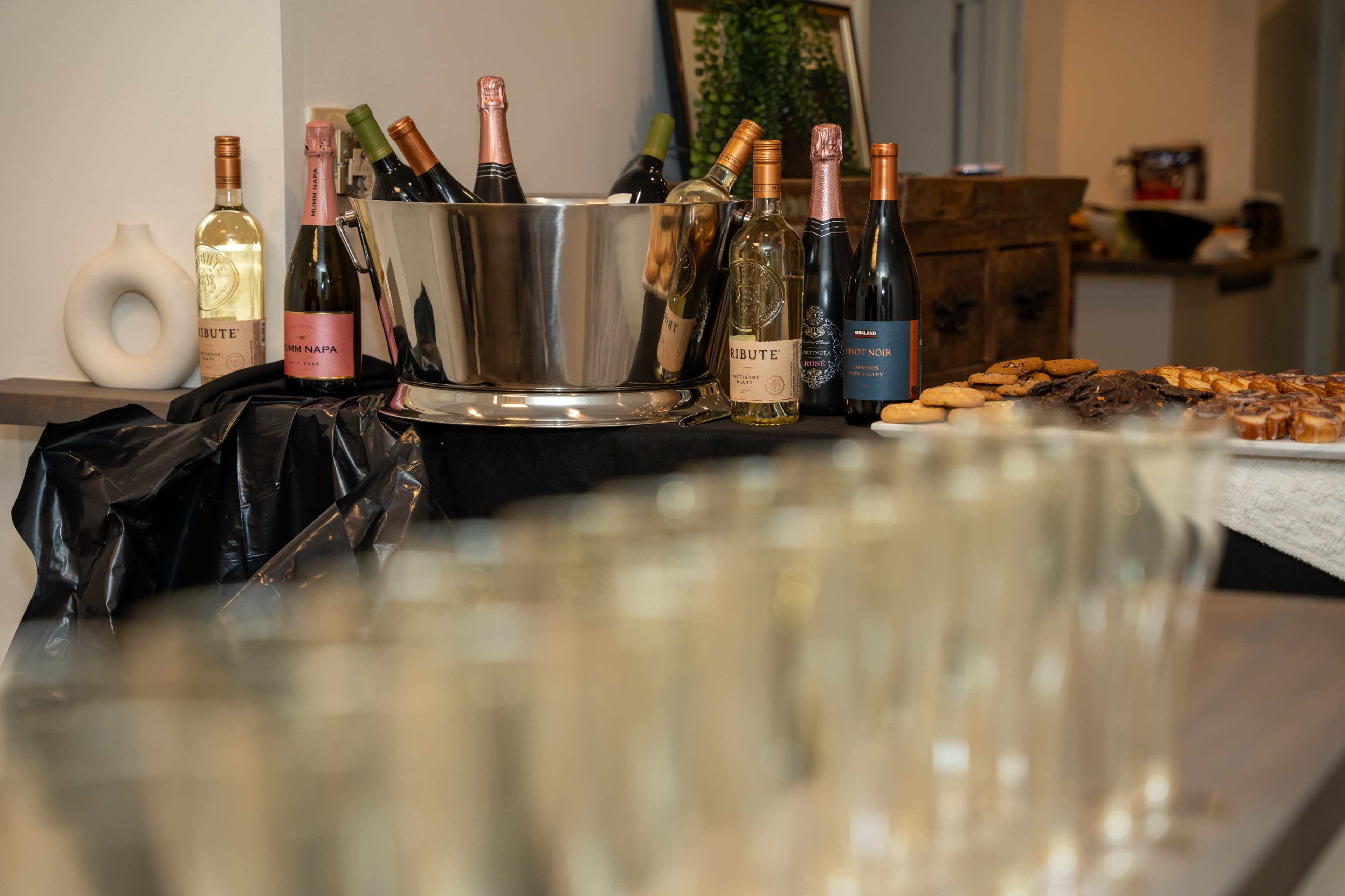 A table is set with a silver ice bucket filled with assorted sparkling wine bottles, alongside a row of clear glasses and plates of pastries.
