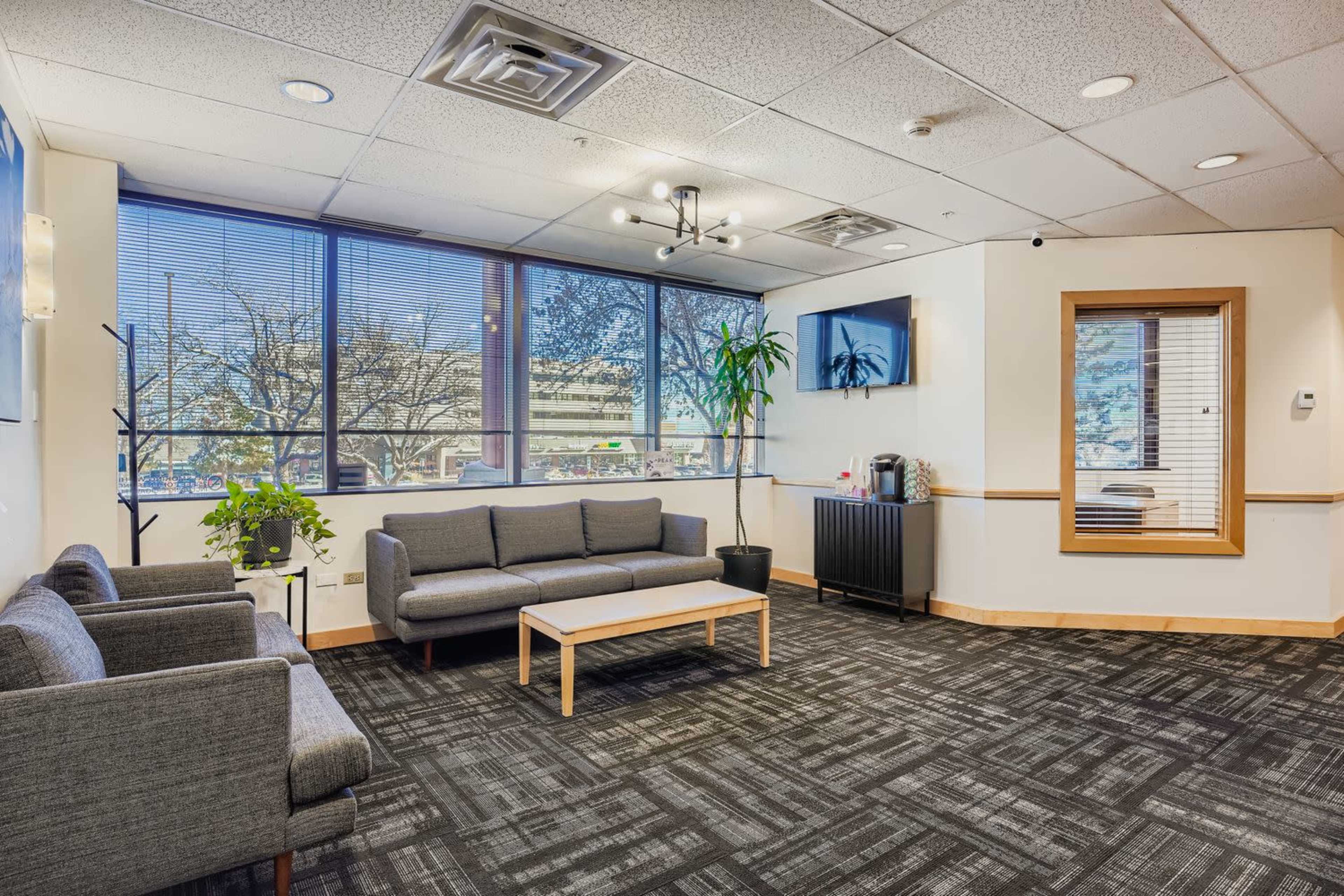 The image shows a waiting area featuring a gray sofa set, a coffee table, and large windows providing natural light with a view of outdoor greenery.