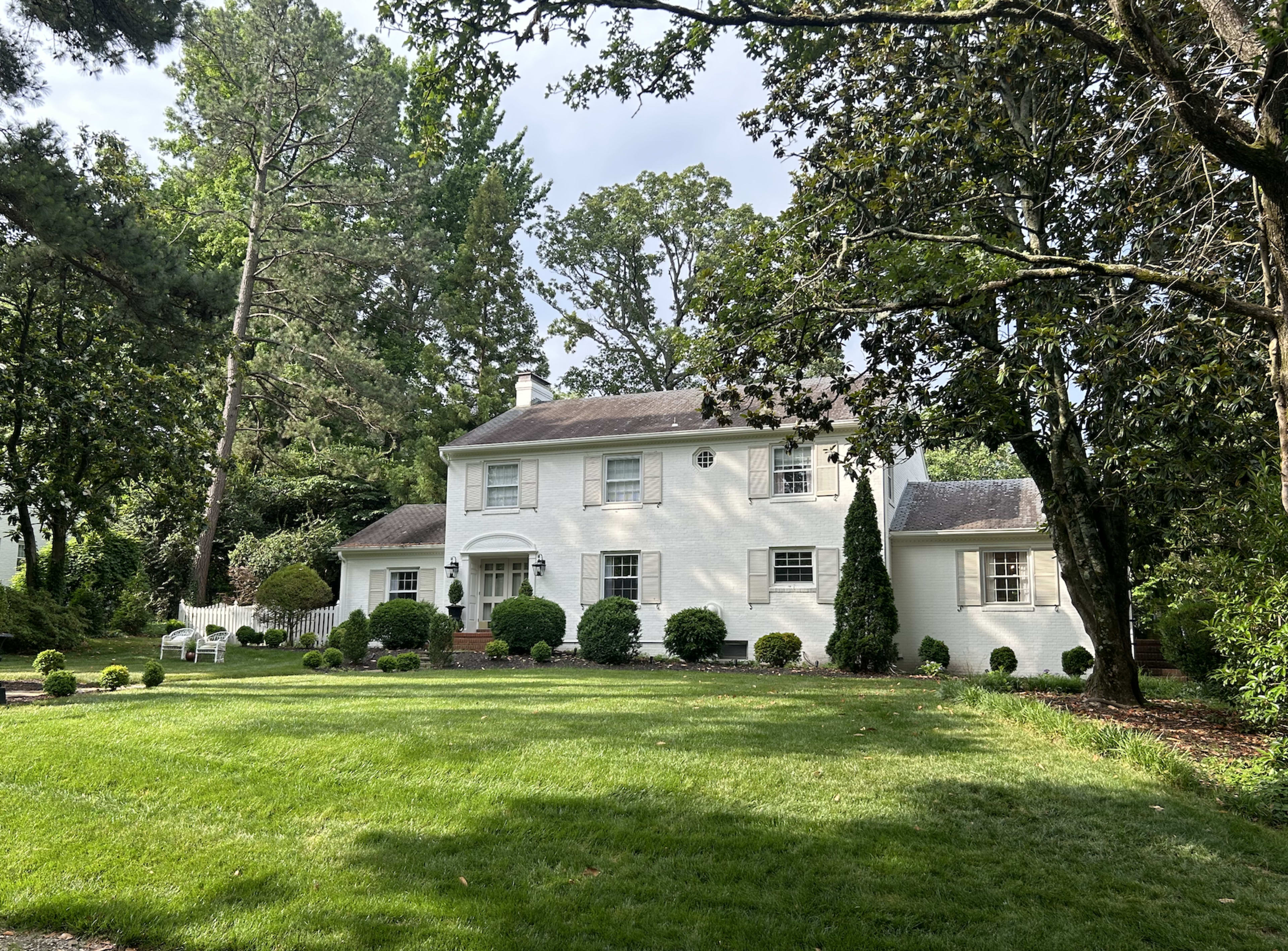 A large white house with a symmetrical facade is surrounded by greenery and well-maintained landscaping.