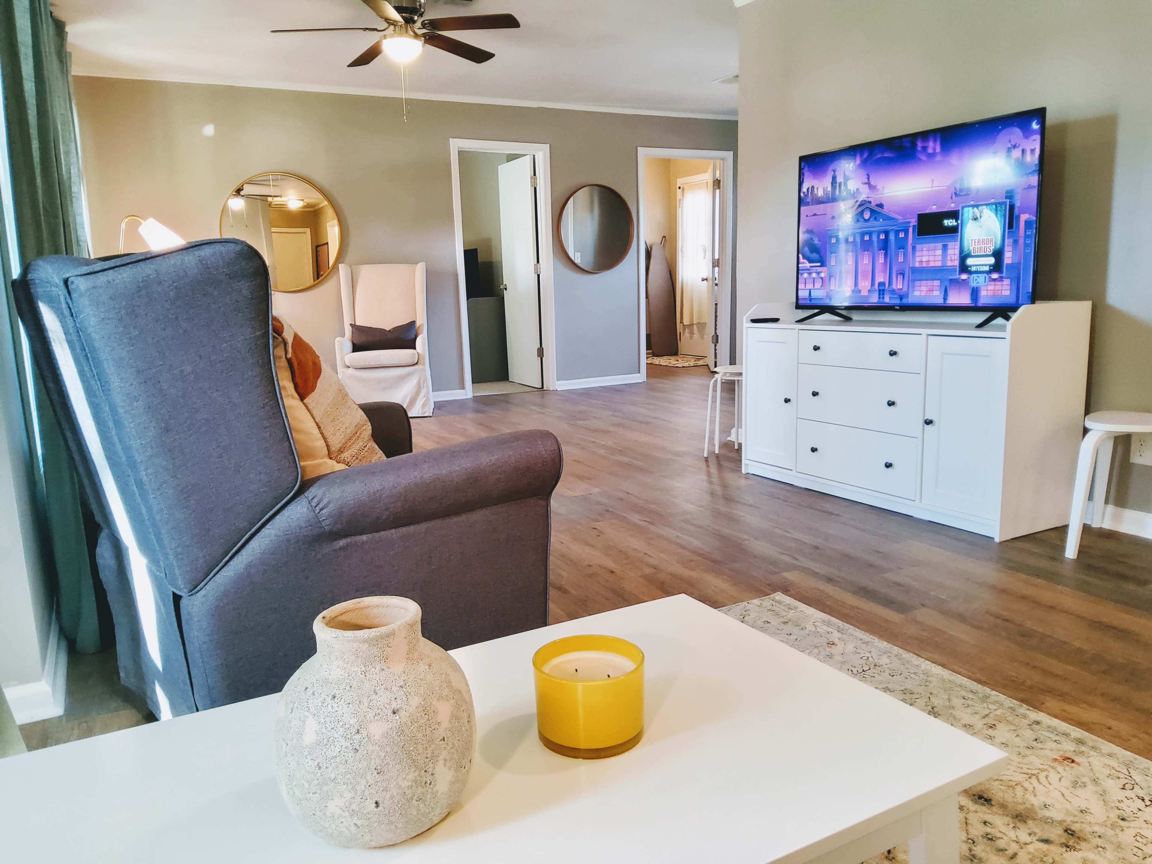 A living room featuring a grey armchair, a coffee table with a candle, a television against the wall, and a door leading to another room.