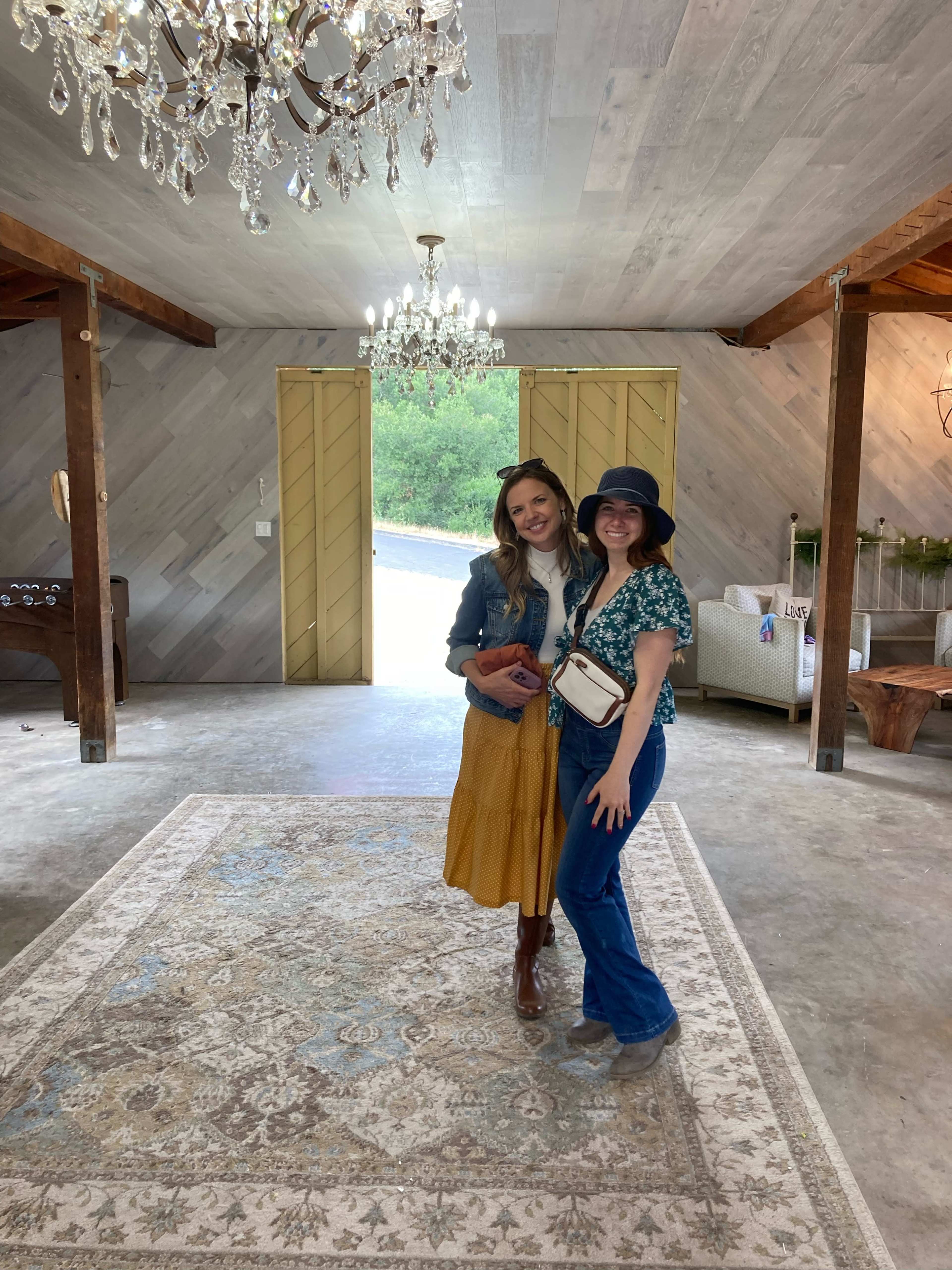 Two women pose together inside a spacious room with wooden beams, chandeliers, and a patterned area rug.