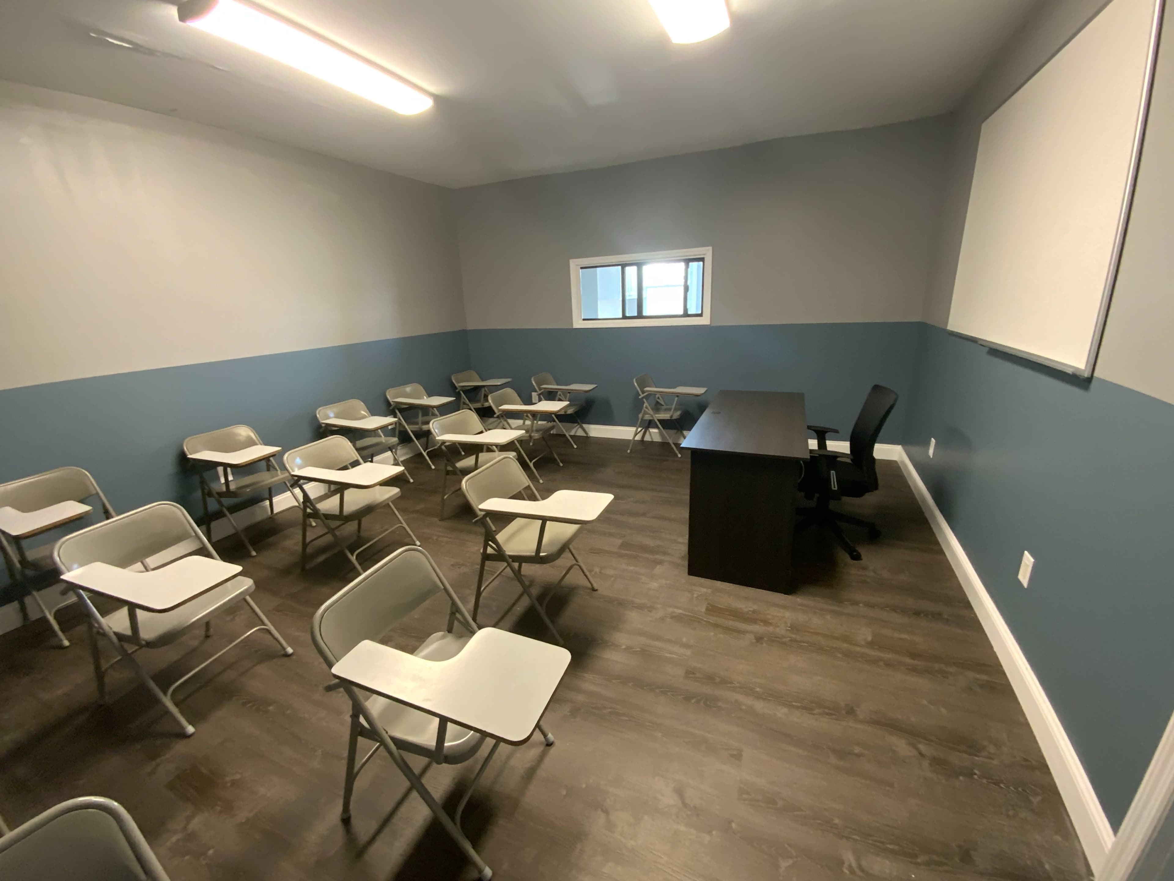 A classroom with empty desks arranged in rows, a black chair, and a desk positioned at the front near a whiteboard.