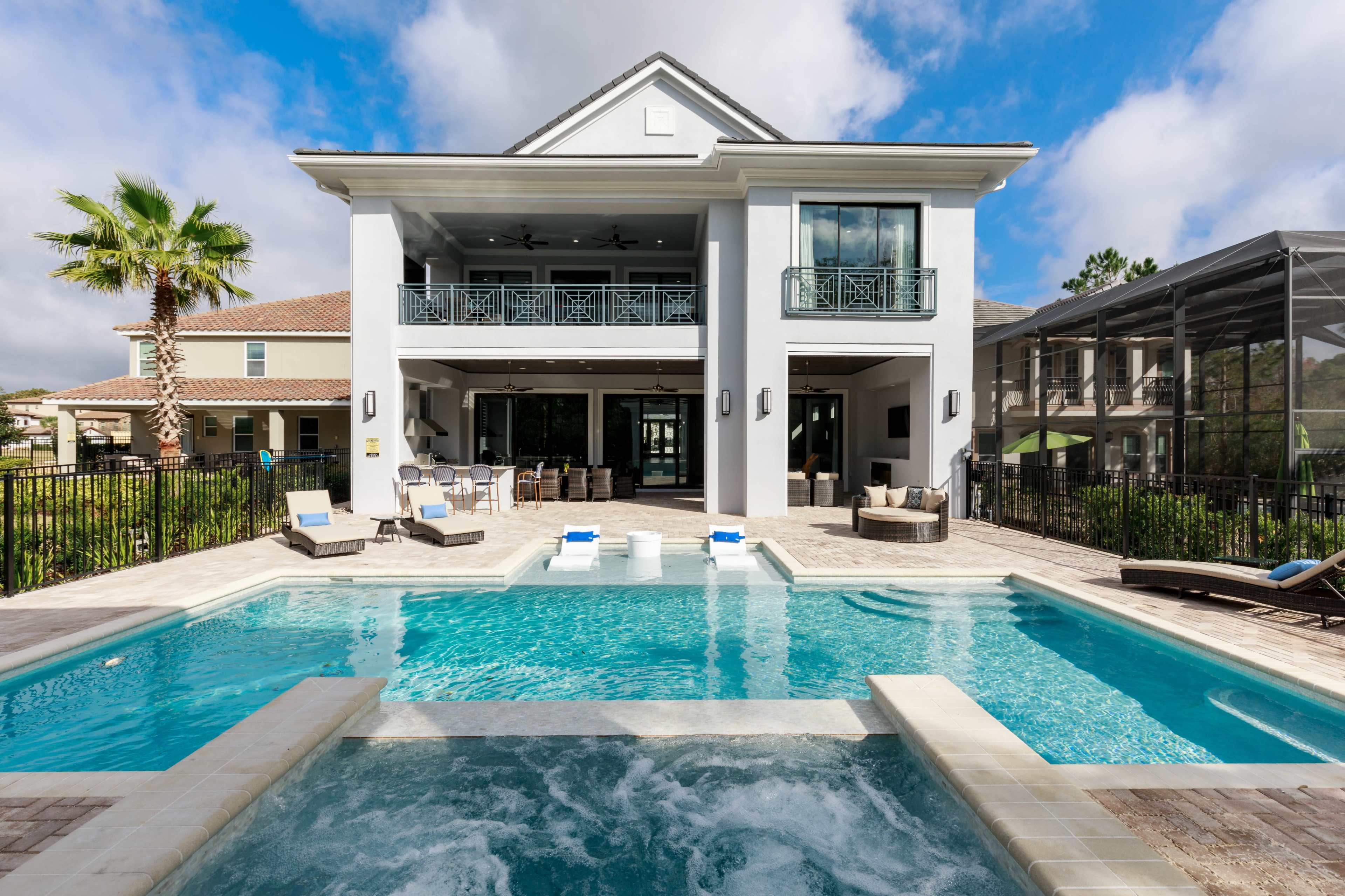 The image shows a modern house with a two-story structure, featuring a spacious pool area with a hot tub, surrounded by a fenced patio and palm trees.