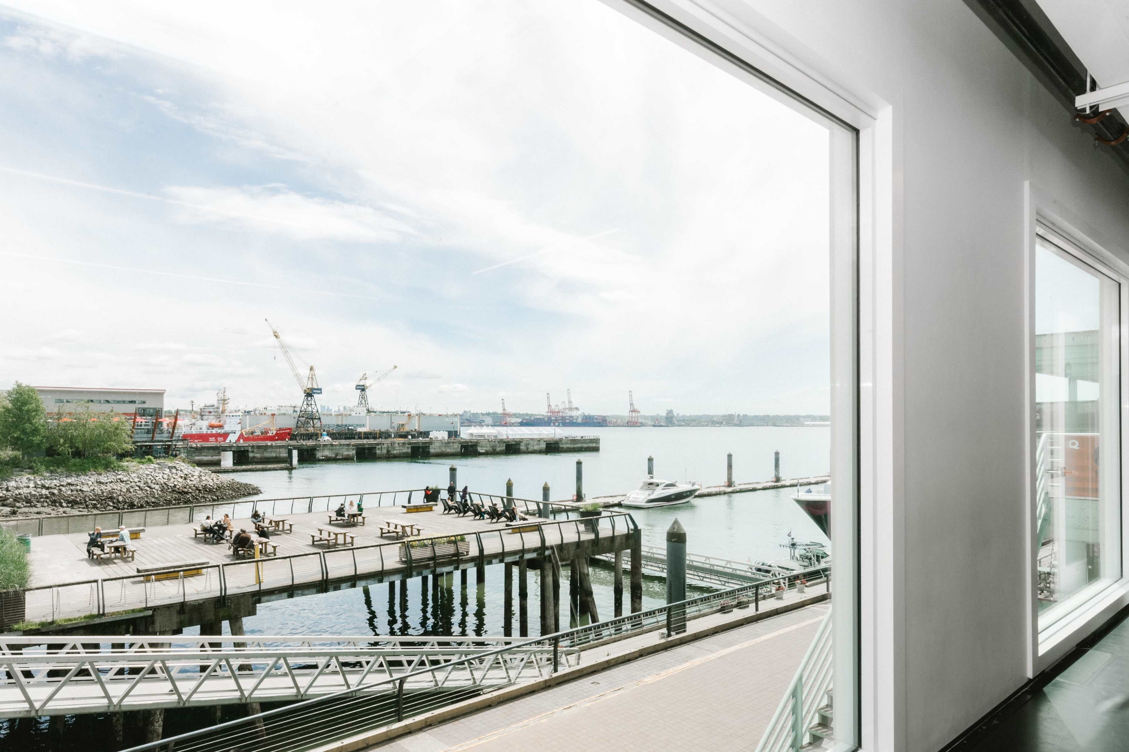 The image shows a view of a harbor with several boats and cranes in the distance, framed by large windows.