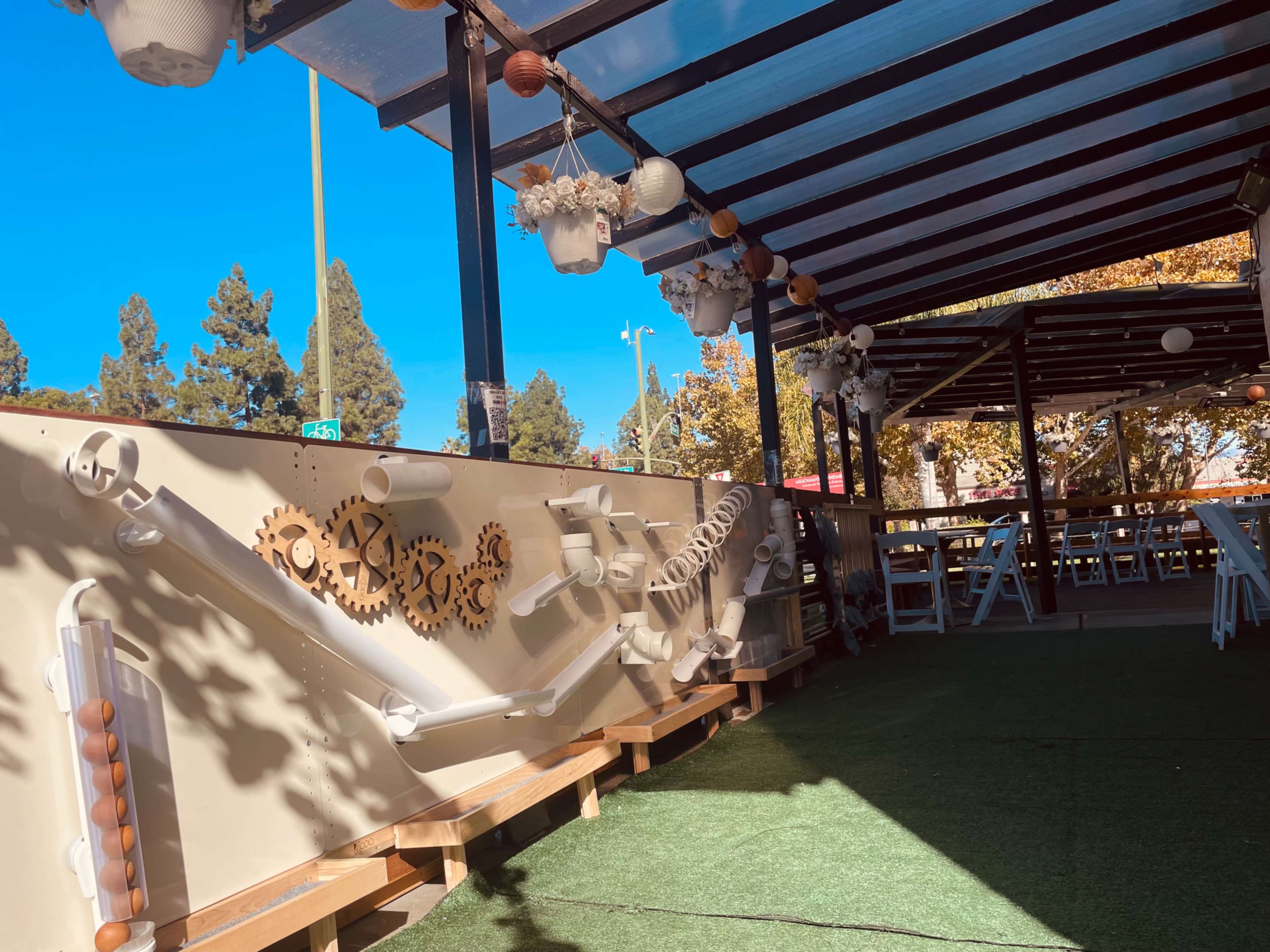 The image shows a playful outdoor space featuring a series of white pipes and gears attached to a wall, with hanging plants and a covered seating area in the background.