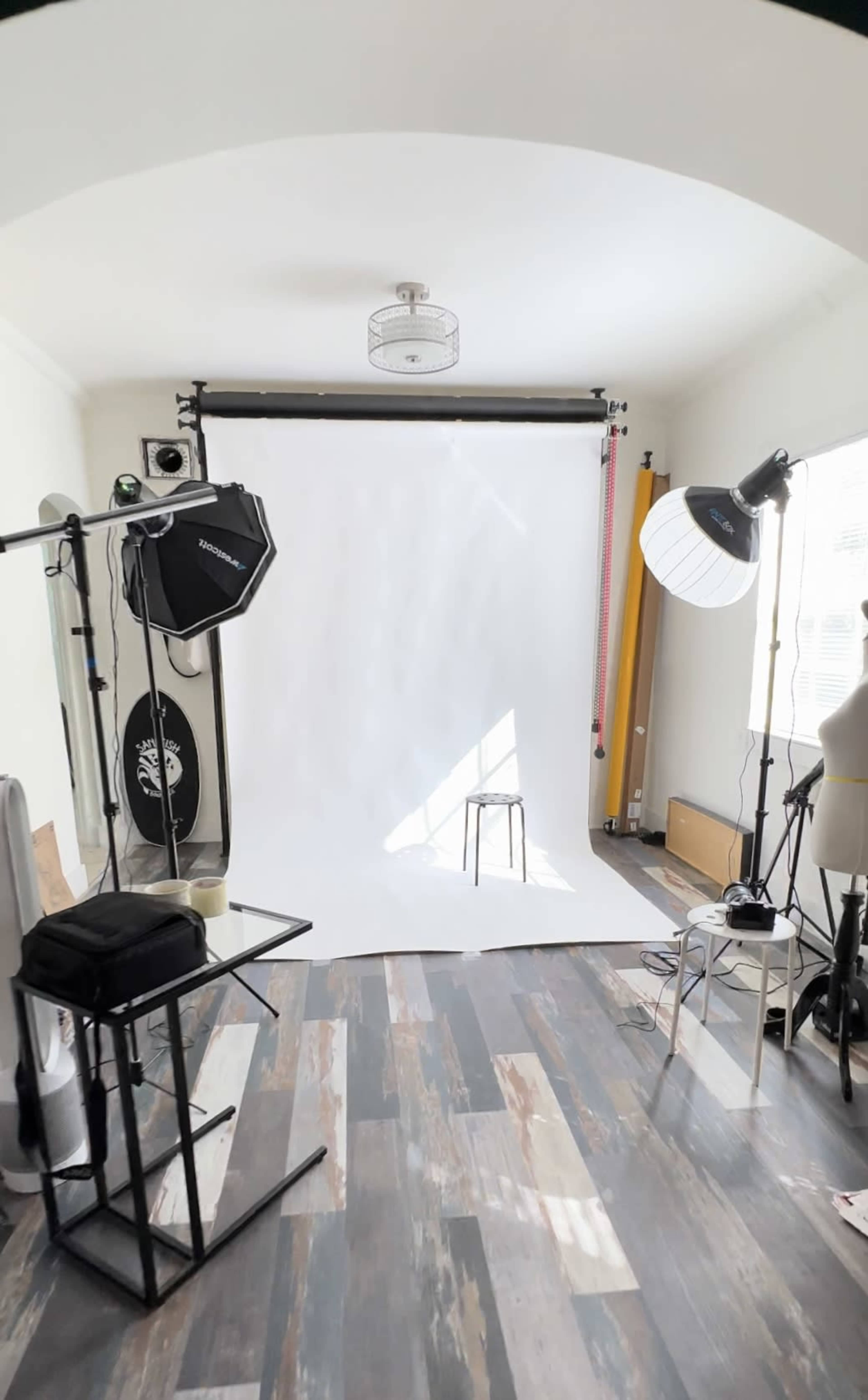 The image shows a photography studio with a white backdrop, various lighting equipment, and a stool in the center.