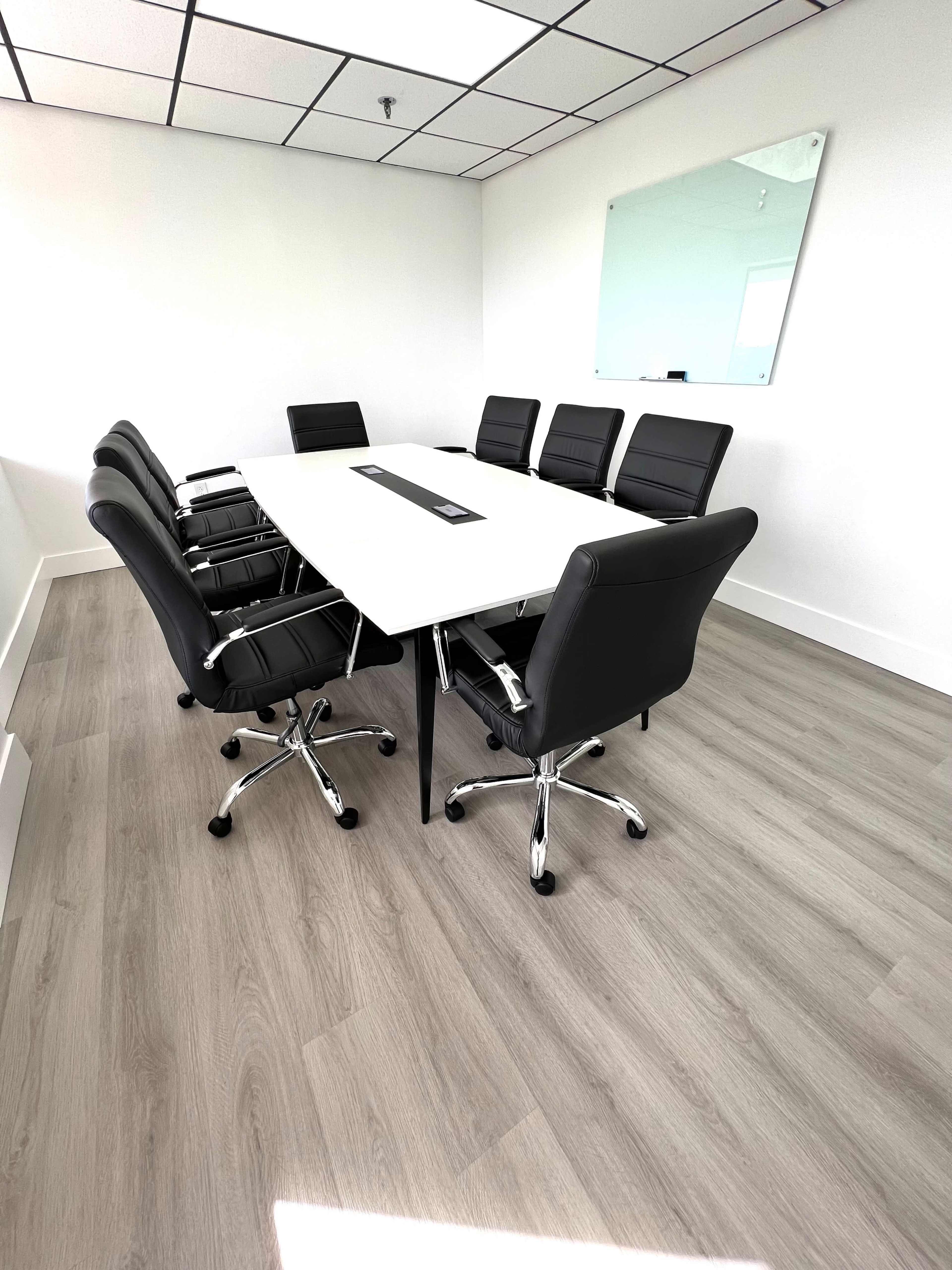 A modern conference room features a rectangular white table surrounded by six black leather chairs, with a large wall mirror reflecting the space.