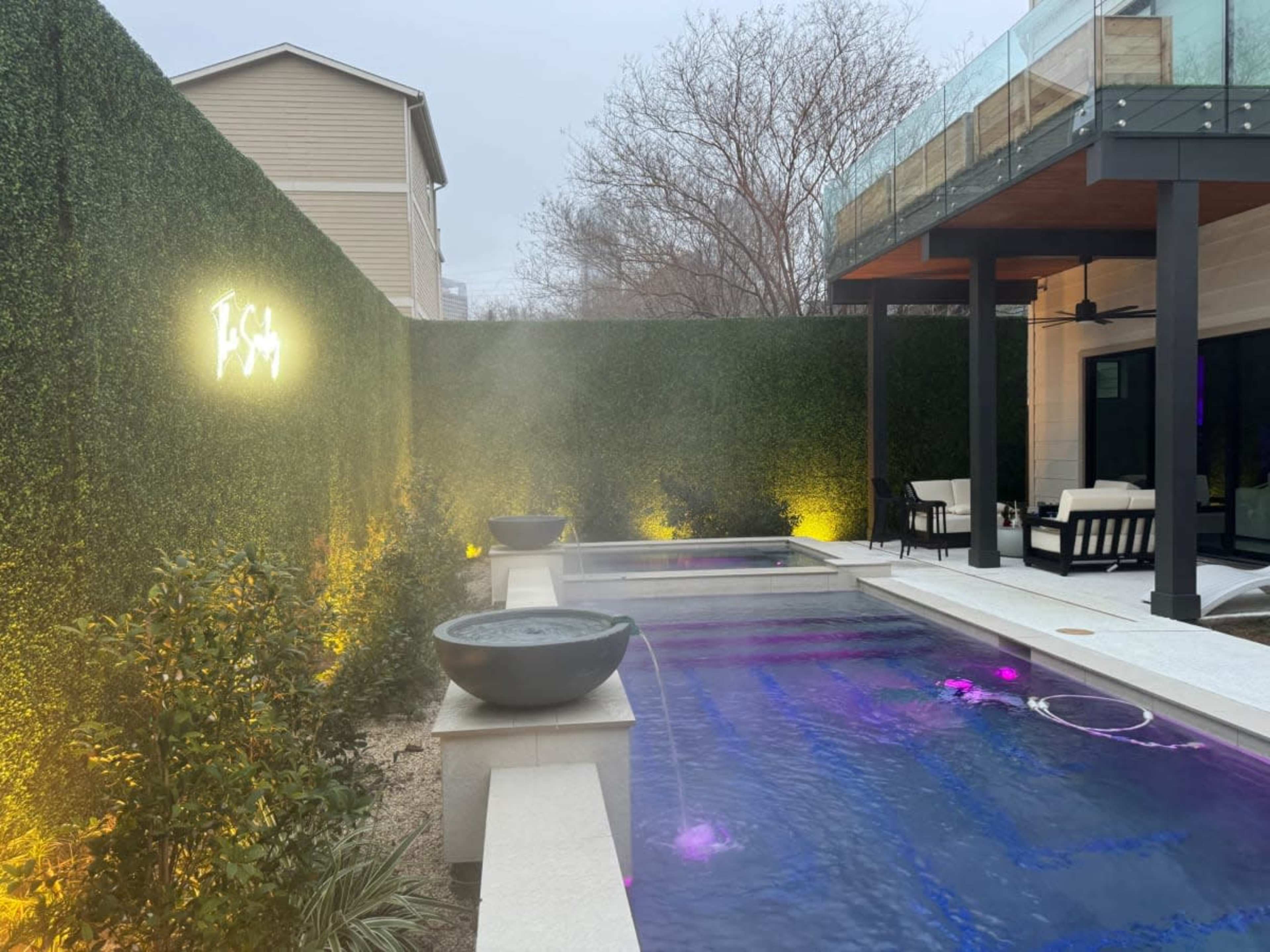 The image shows a modern pool area with stone borders, illuminated water features, and a backdrop of tall greenery and a covered seating area.