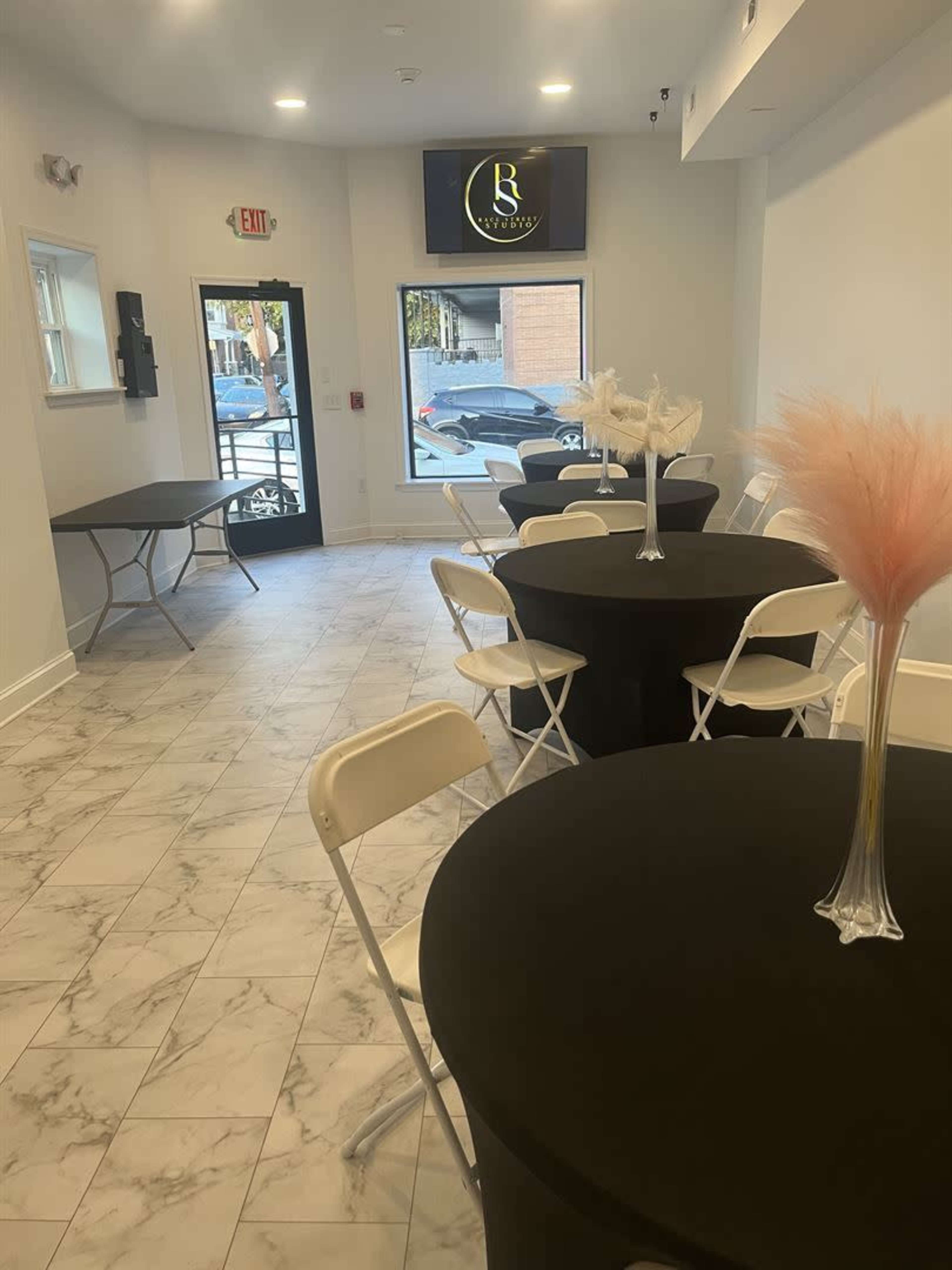 The image shows a white-walled event space with marble flooring, featuring several round black tables set with vases and tall decorative plants, along with a large screen displaying a logo on the wall.