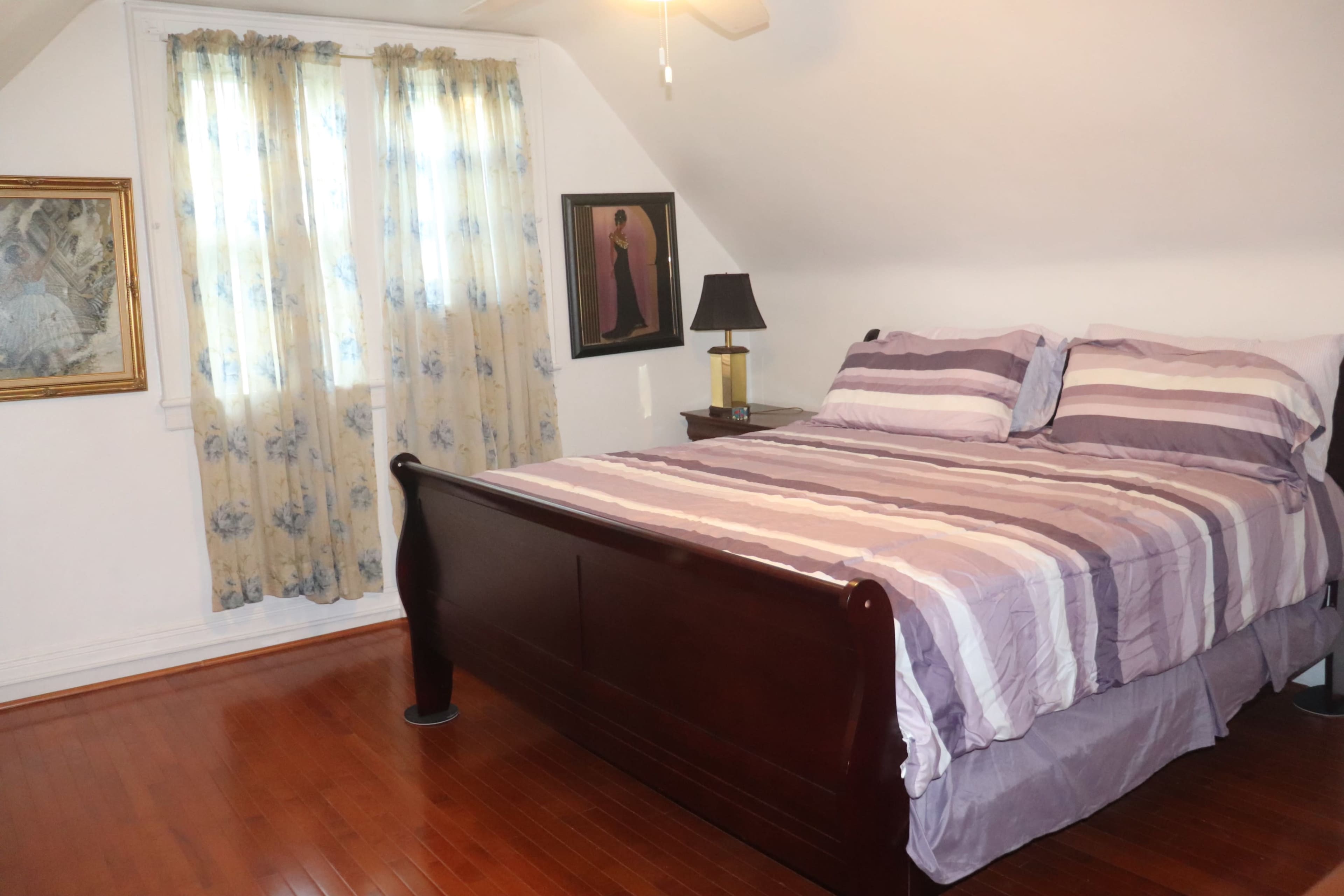 A bedroom with a wooden sleigh bed, striped bedding, a bedside lamp, and two windows covered with light curtains.