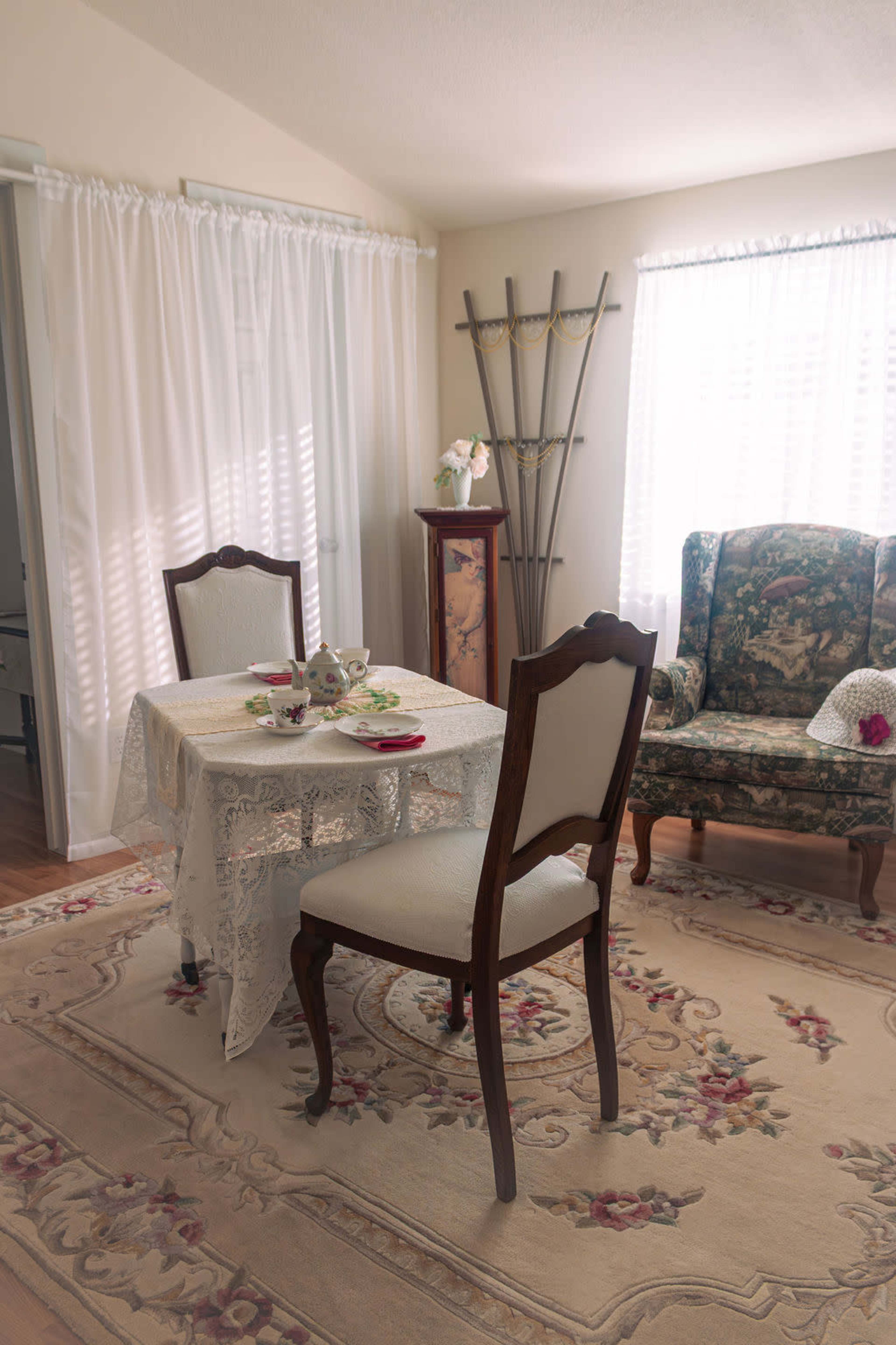 A dining area features a lace tablecloth set with tea cups and a single empty chair beside a patterned armchair.