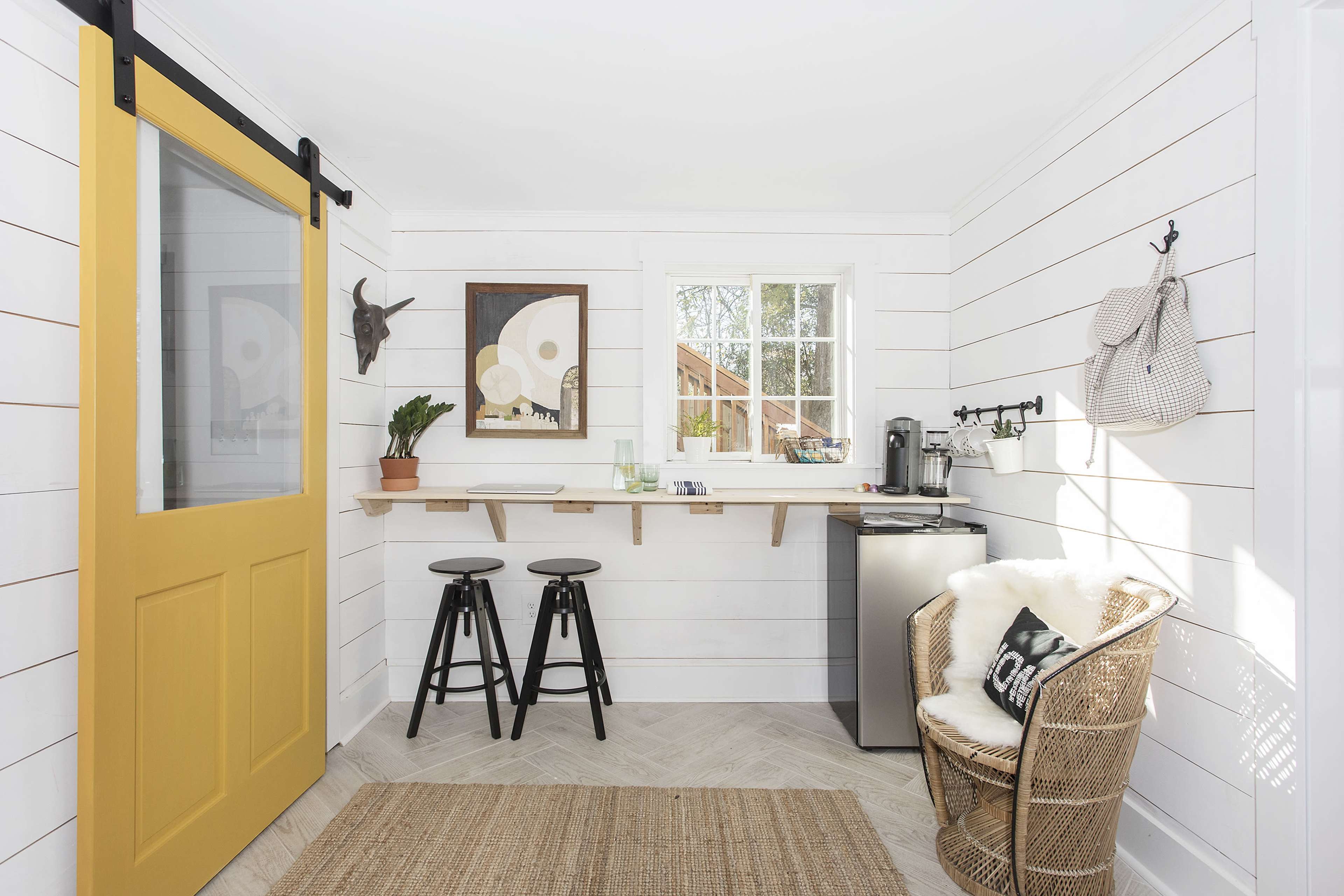 A small, bright room featuring a yellow sliding door, a wooden shelf, two black stools, a kitchenette with a mini fridge, and a rattan chair.