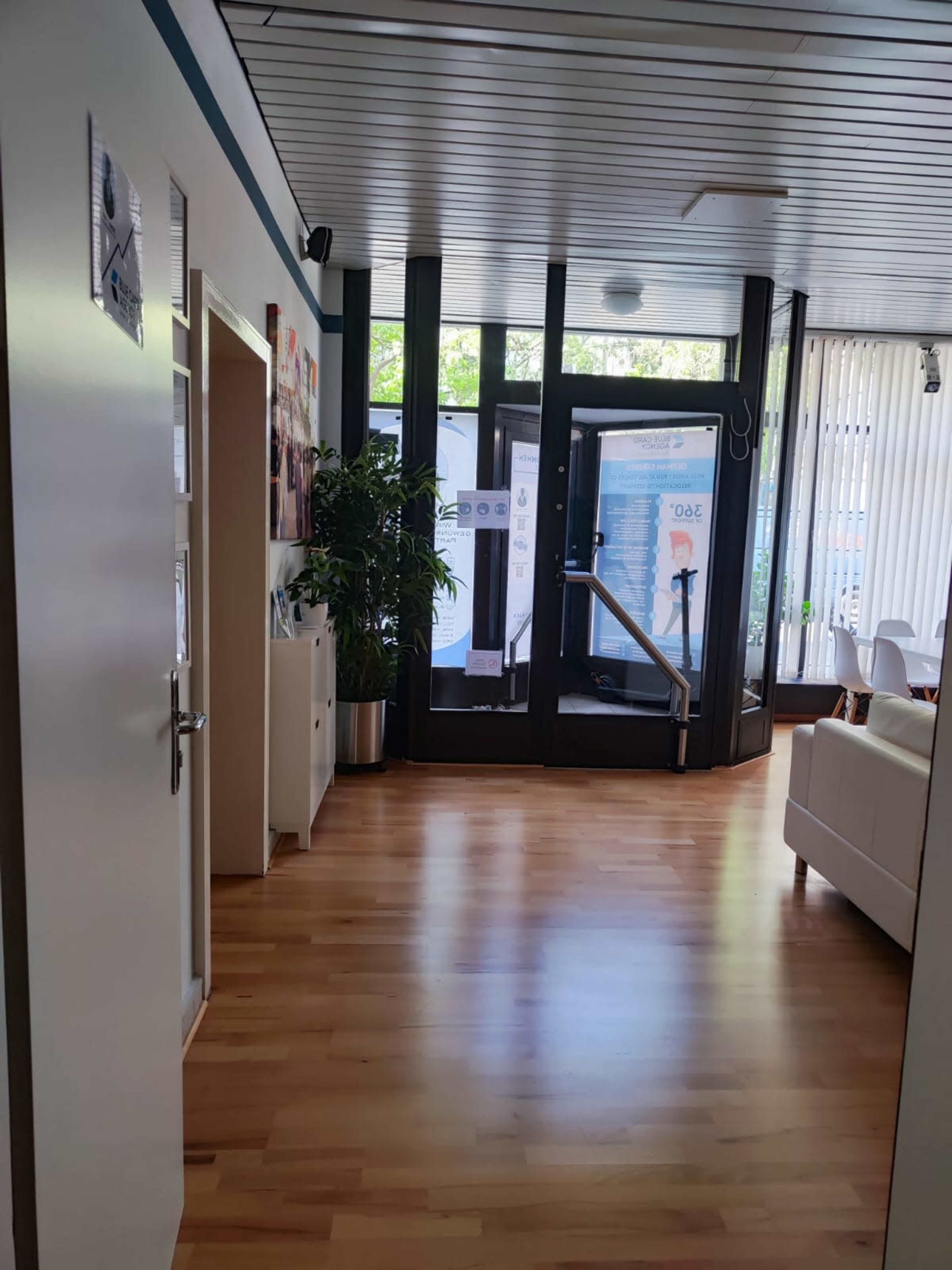 The image shows a modern indoor hallway leading to a glass entrance with wooden flooring and bright overhead lighting.