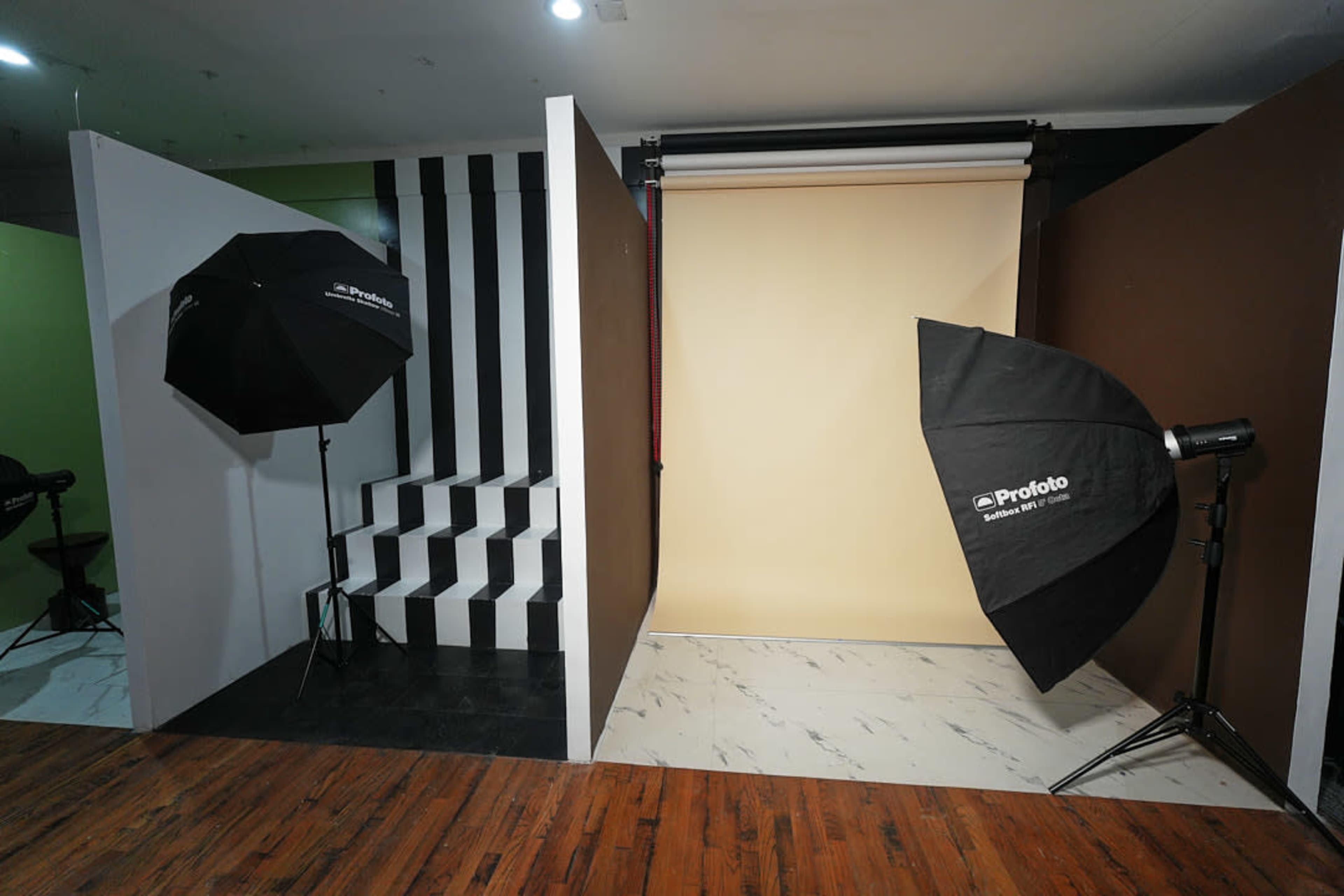The image shows a photography studio setup with two umbrella softboxes, a staircase with black and white stripes, and a beige backdrop on one side.