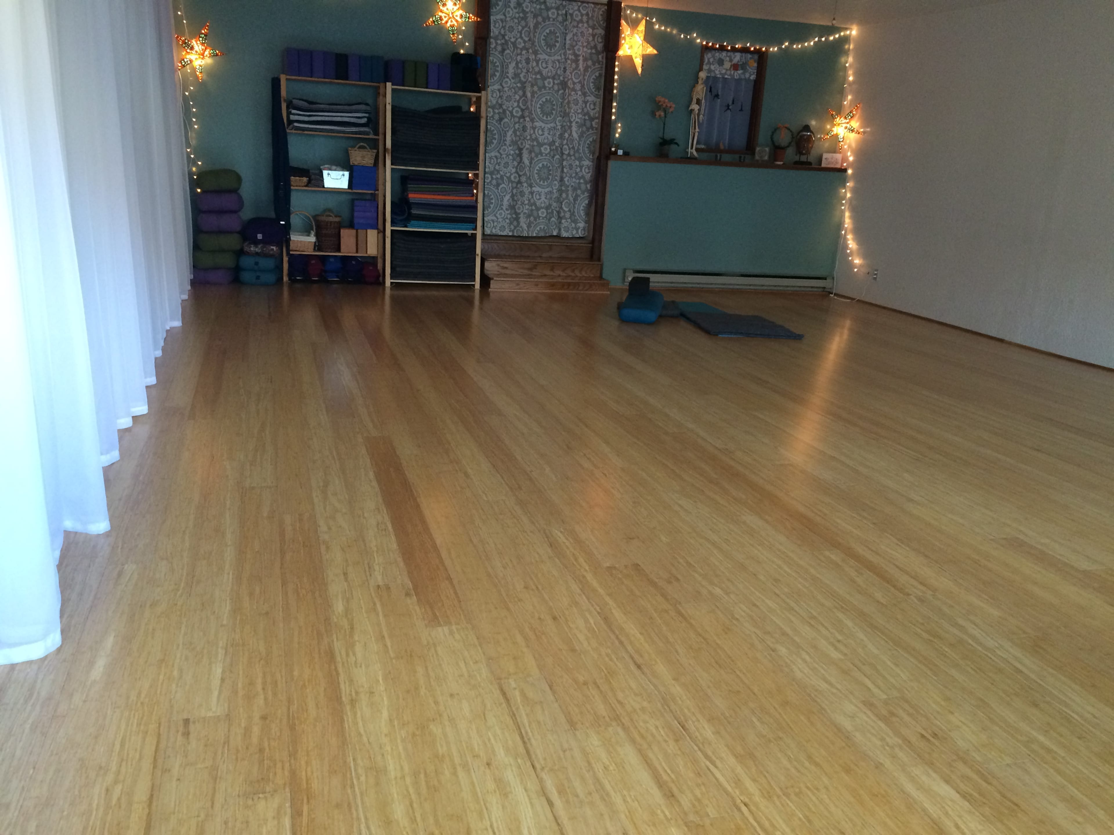 The image shows a spacious yoga studio with bamboo flooring, shelves holding yoga props, and soft lighting in a calm atmosphere.