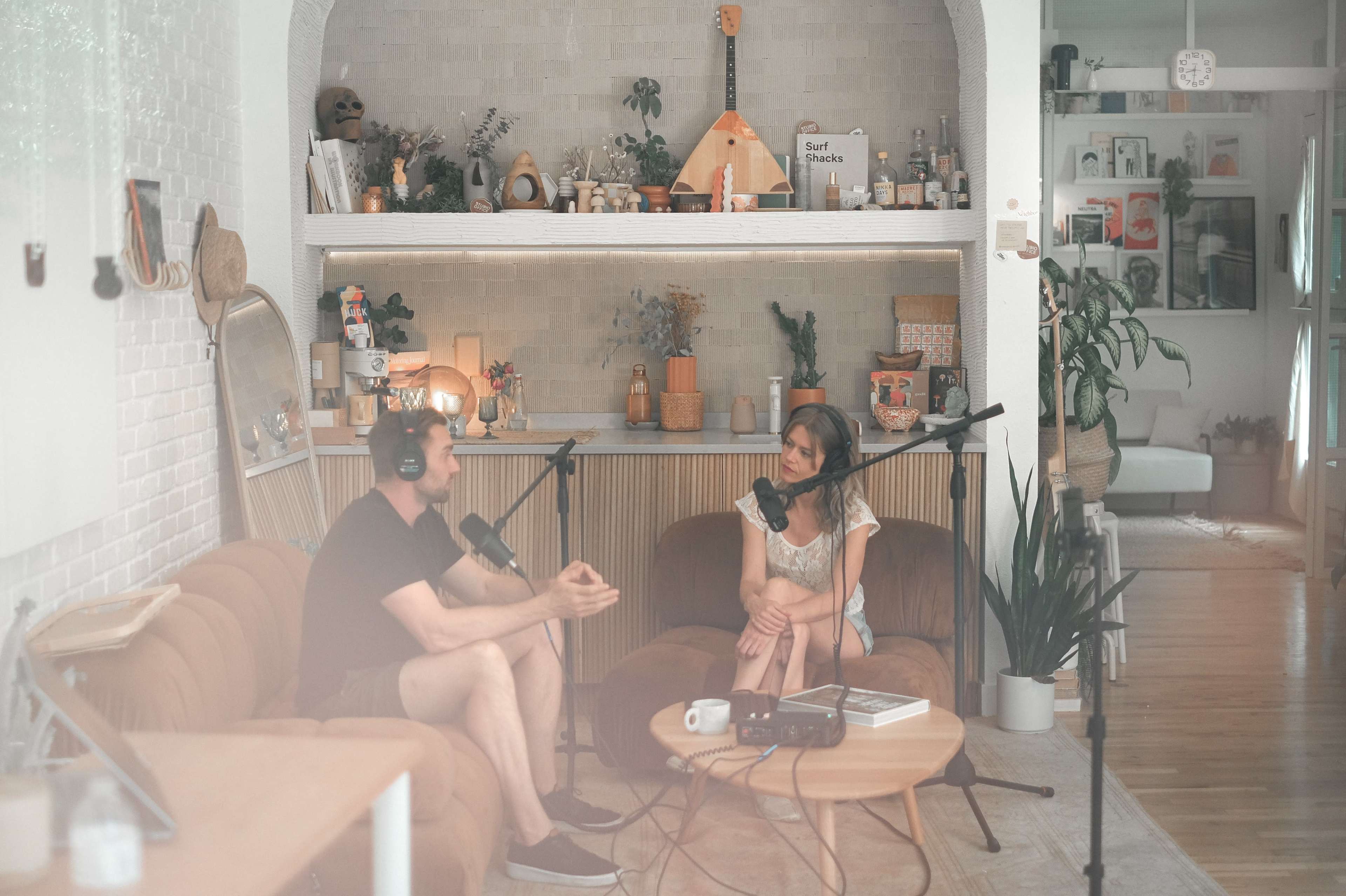 A man and a woman are engaged in a conversation in a cozy, well-decorated podcast studio with microphones and cameras set up.