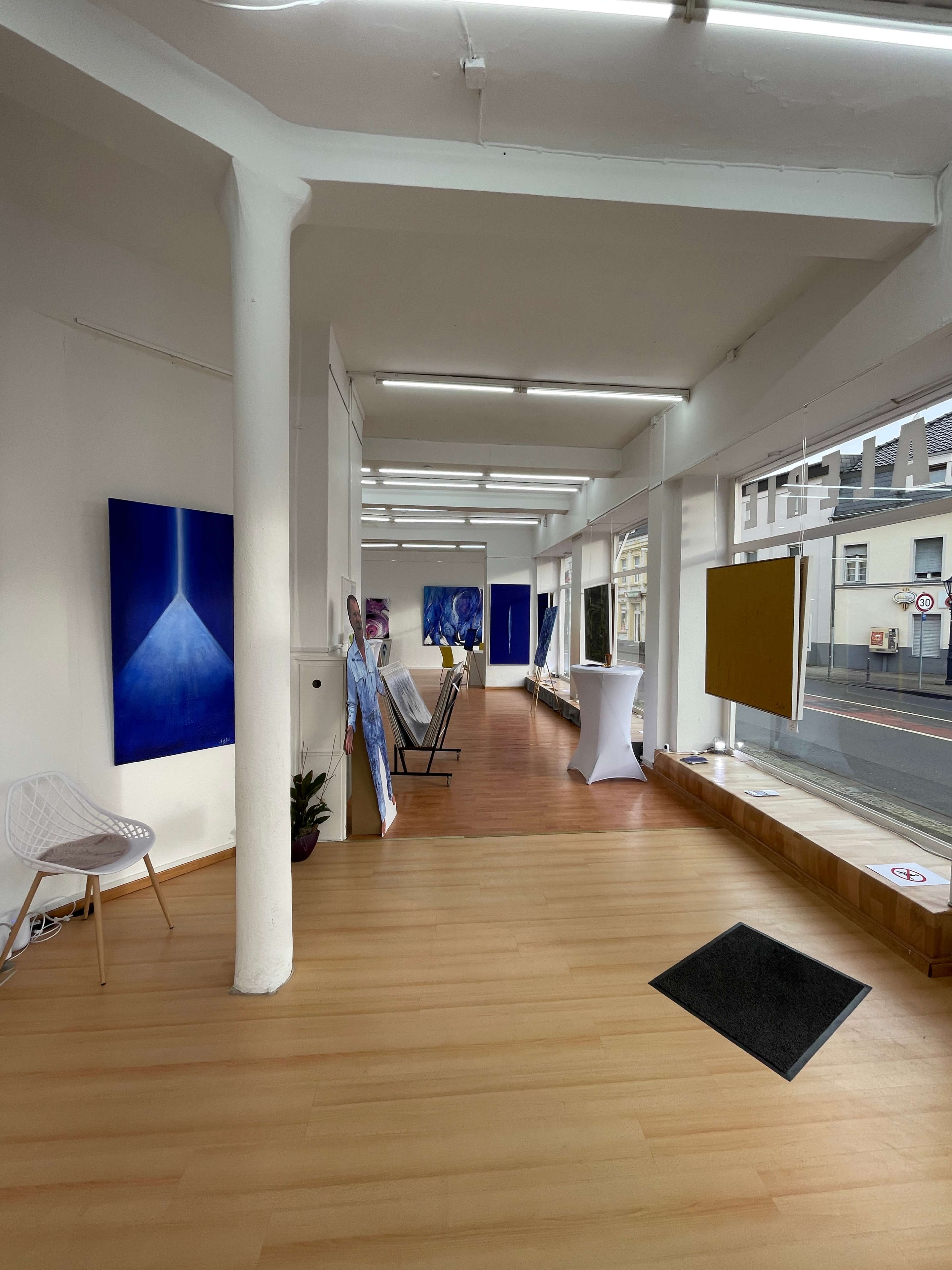 An art gallery with wooden flooring, featuring several paintings displayed along the white walls and a small seating area on the left.