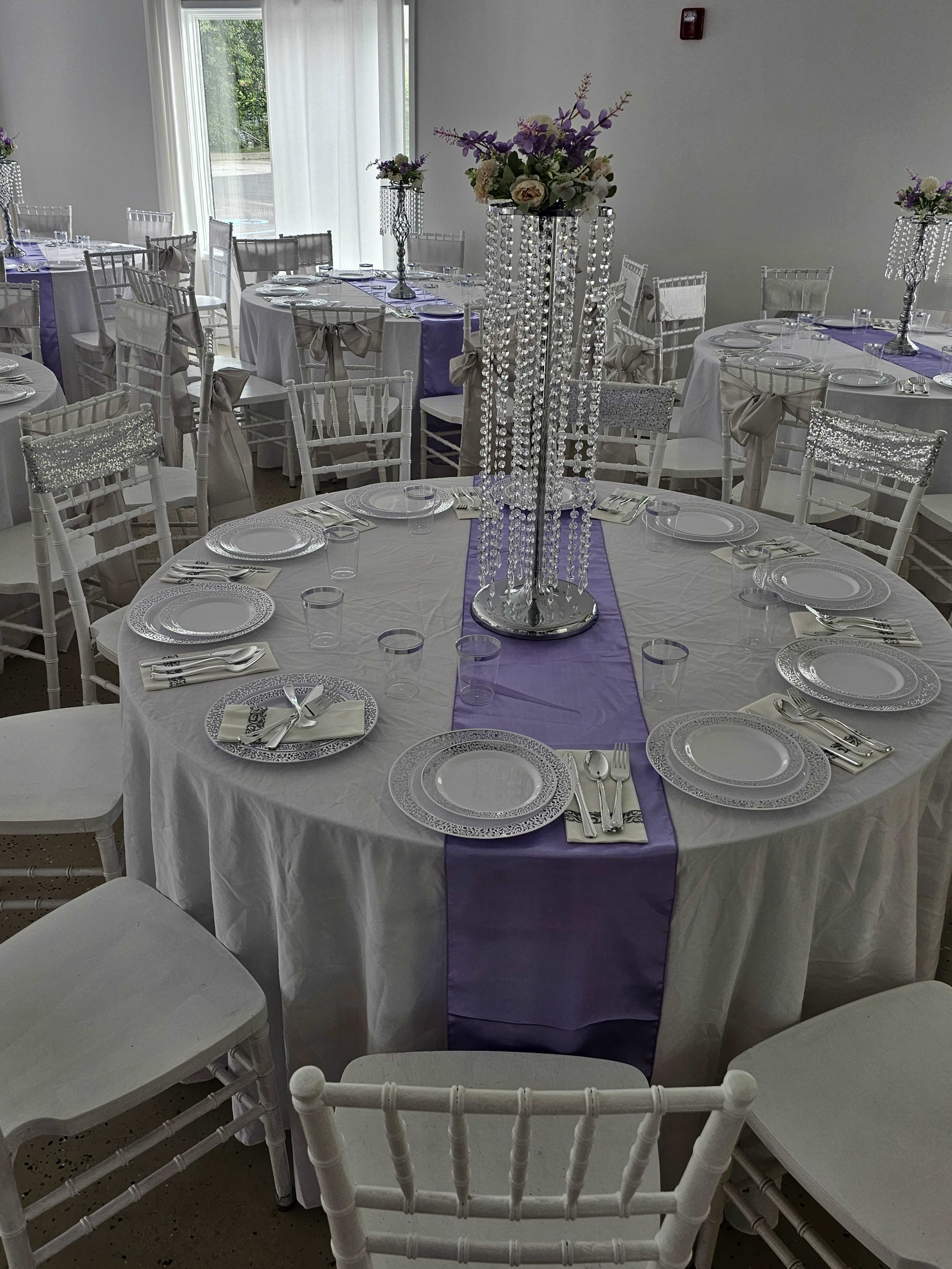 A banquet hall set for a formal event, featuring round tables covered with white tablecloths and lavender runners, adorned with crystal centerpieces and floral arrangements.