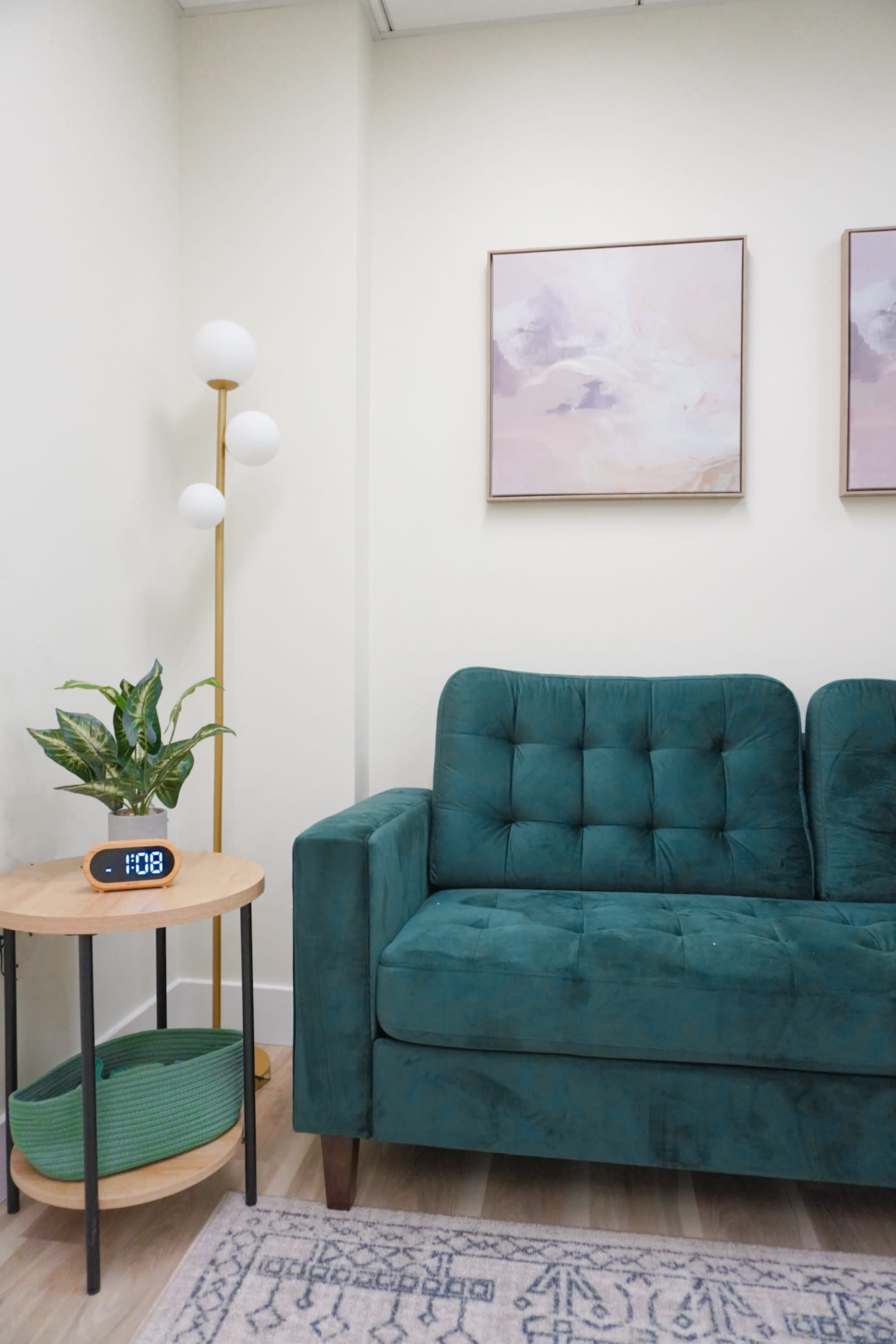 A green tufted sofa sits beside a round wooden table with a plant and a digital clock, against a softly painted wall with framed artwork.