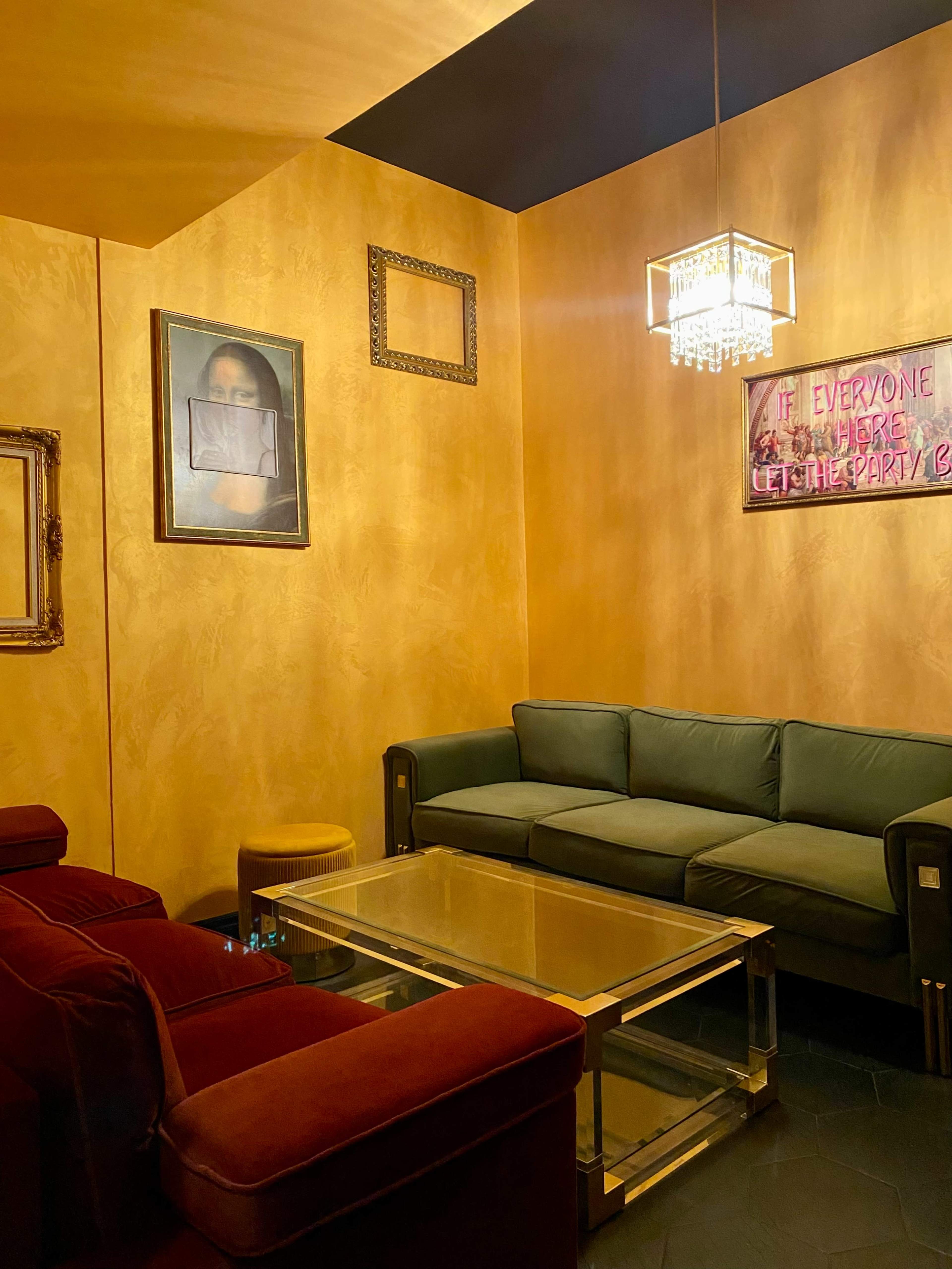 A cozy lounge area with a green couch, red armchairs, and a glass coffee table, illuminated by a chandelier and adorned with framed artwork on golden walls.