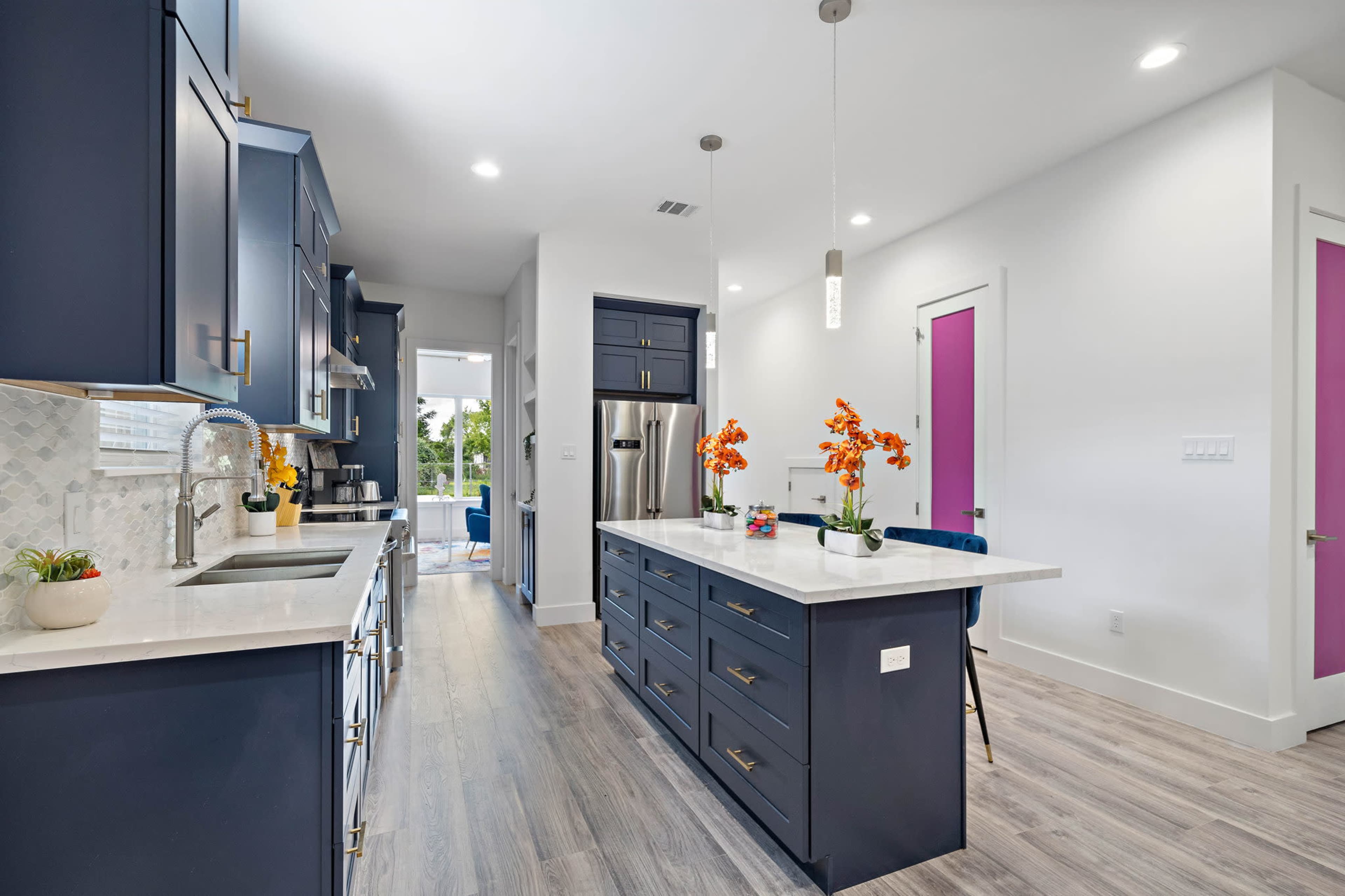 A modern kitchen with dark blue cabinetry, a central island with a white countertop, and a light-filled space featuring vibrant purple doors.