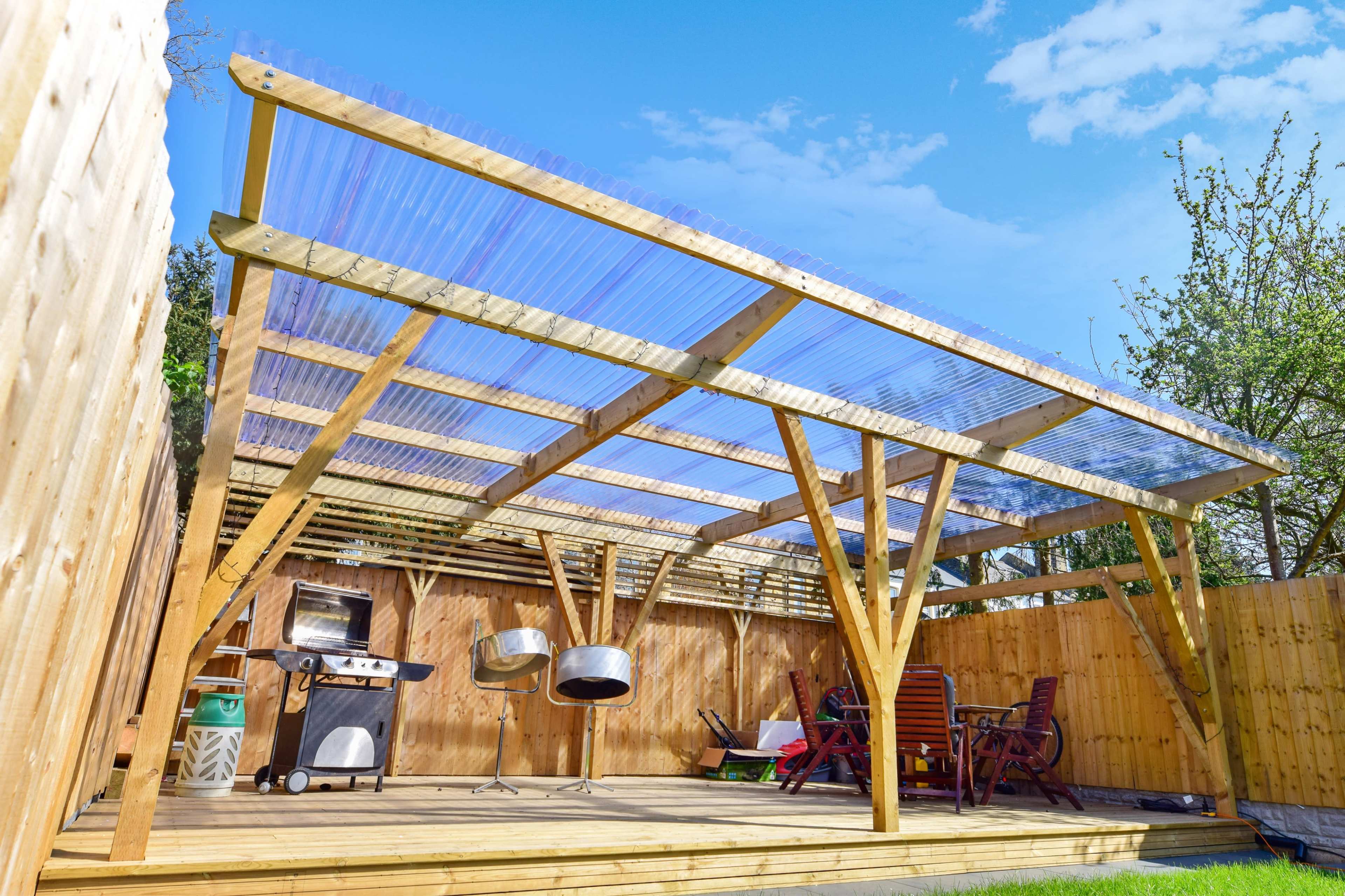 A wooden patio structure with a transparent roof, equipped with a barbecue grill and outdoor seating, situated in a backyard surrounded by wooden fencing.