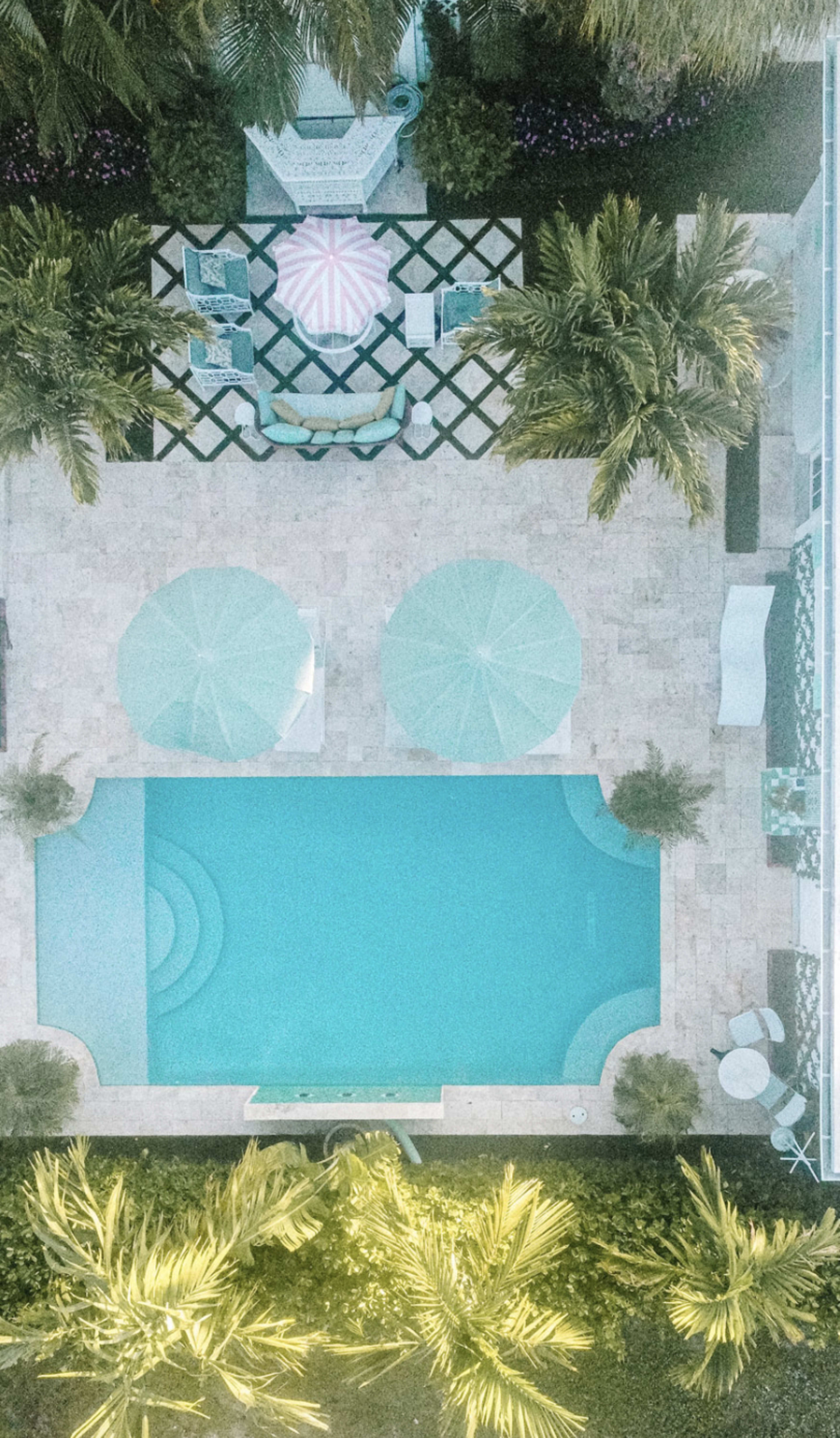 An aerial view shows a swimming pool surrounded by patio furniture and palm trees, with two umbrellas providing shade.