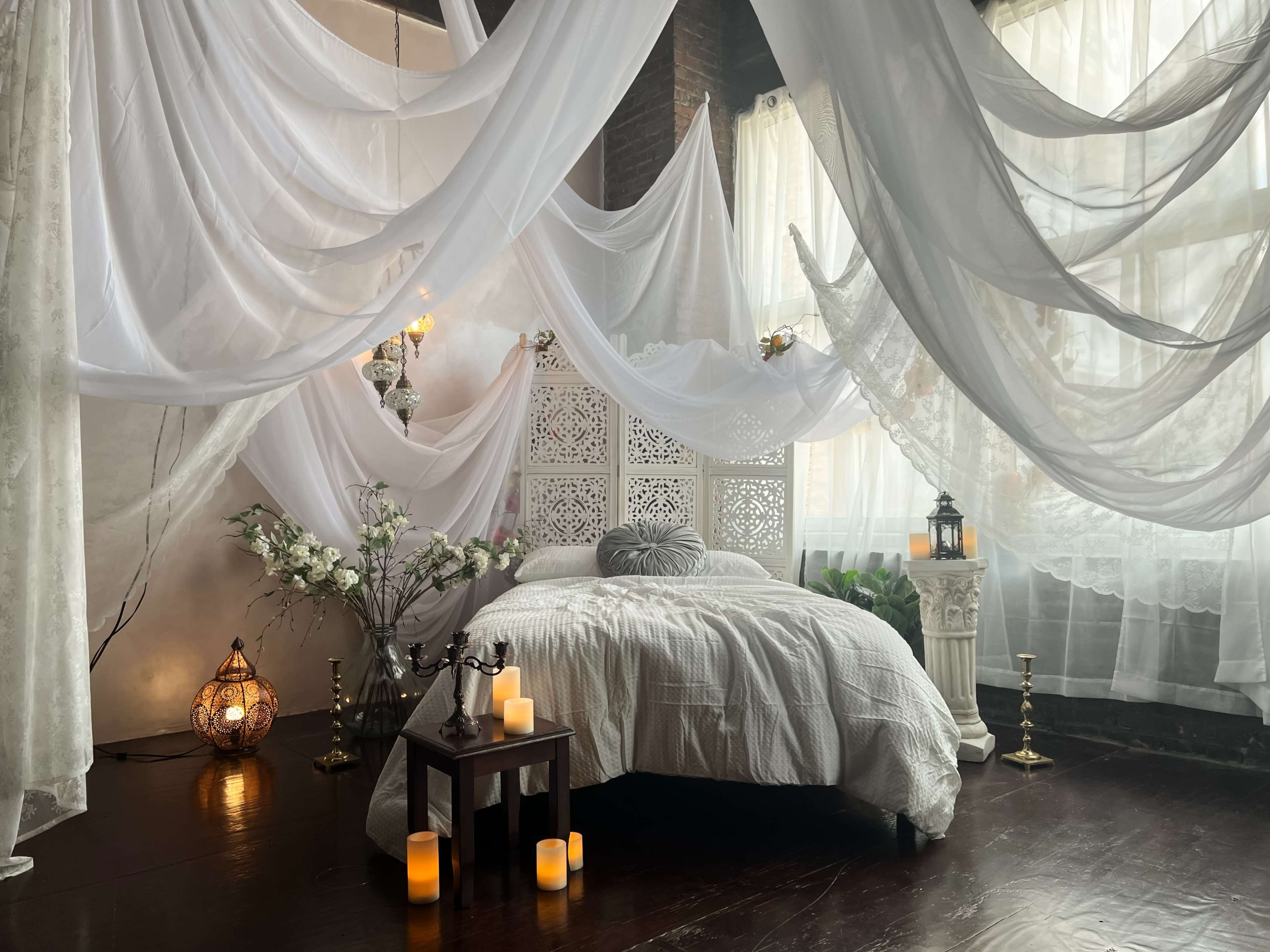 A serene bedroom adorned with flowing white drapes and a neatly made bed surrounded by decorative elements like candles and floral arrangements.