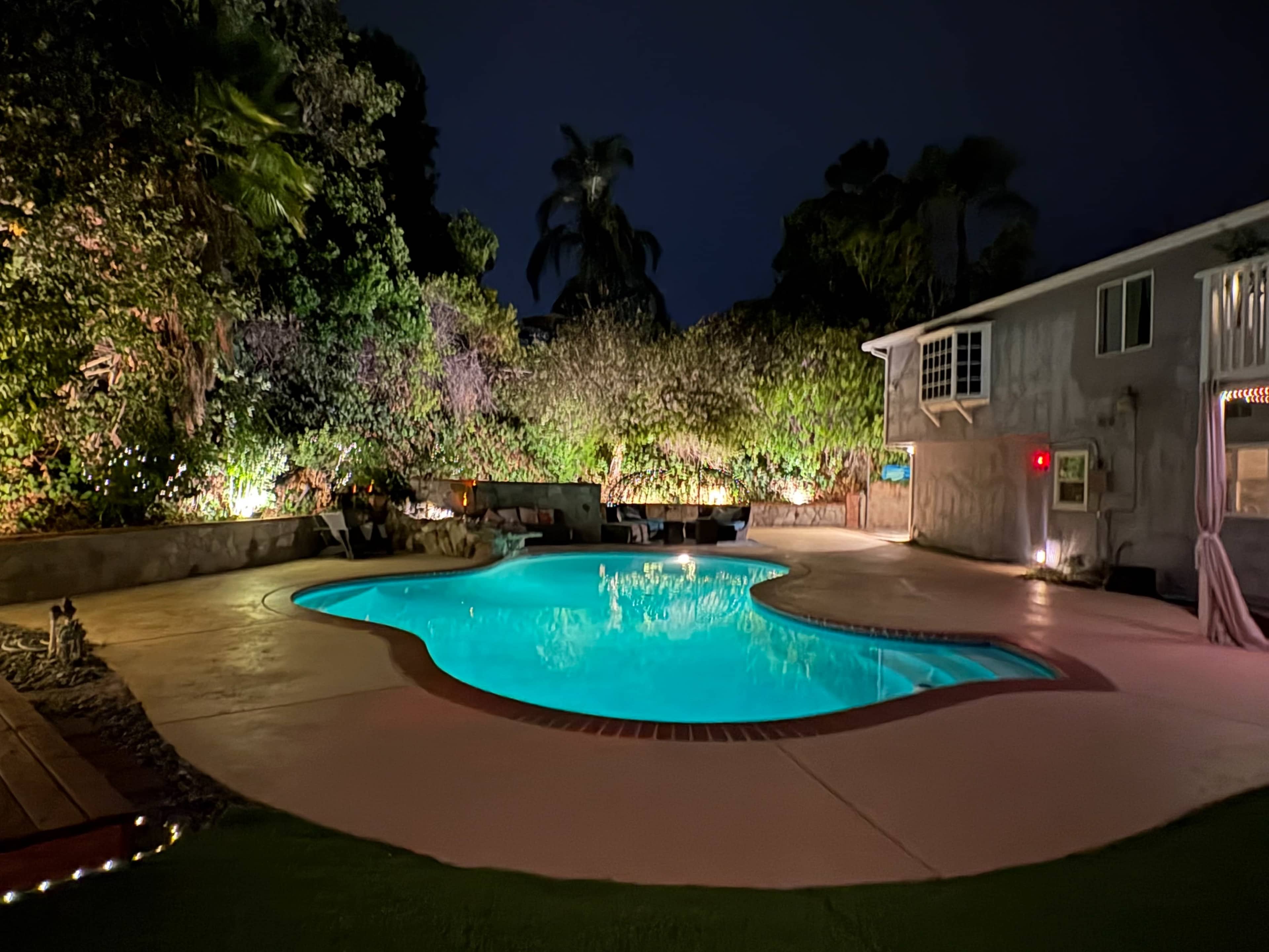 The image shows a backyard pool area at night, illuminated by soft lights, surrounded by lush greenery and a house to one side.