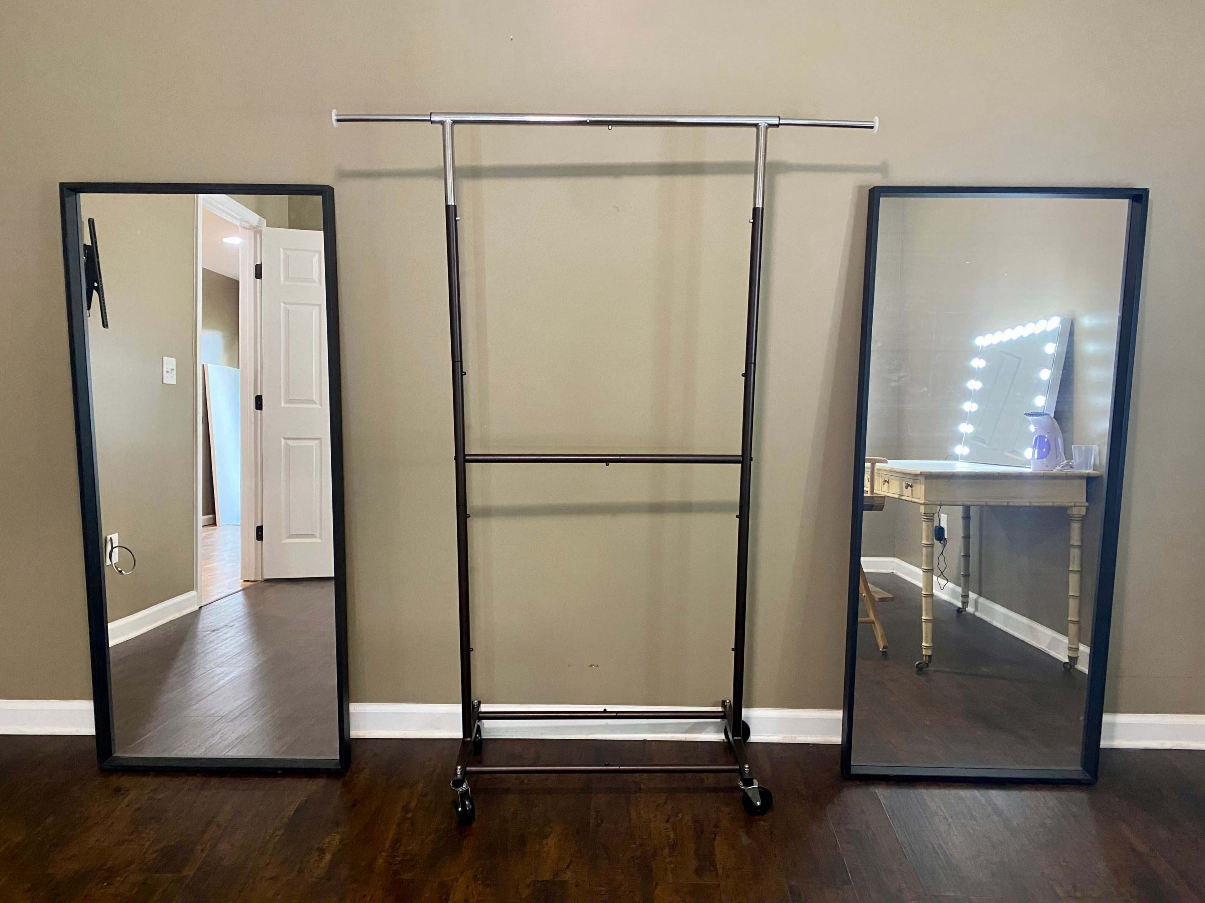 The image shows a clothing rack positioned between two large mirrors in a room with a beige wall and a lighted vanity table in the background.