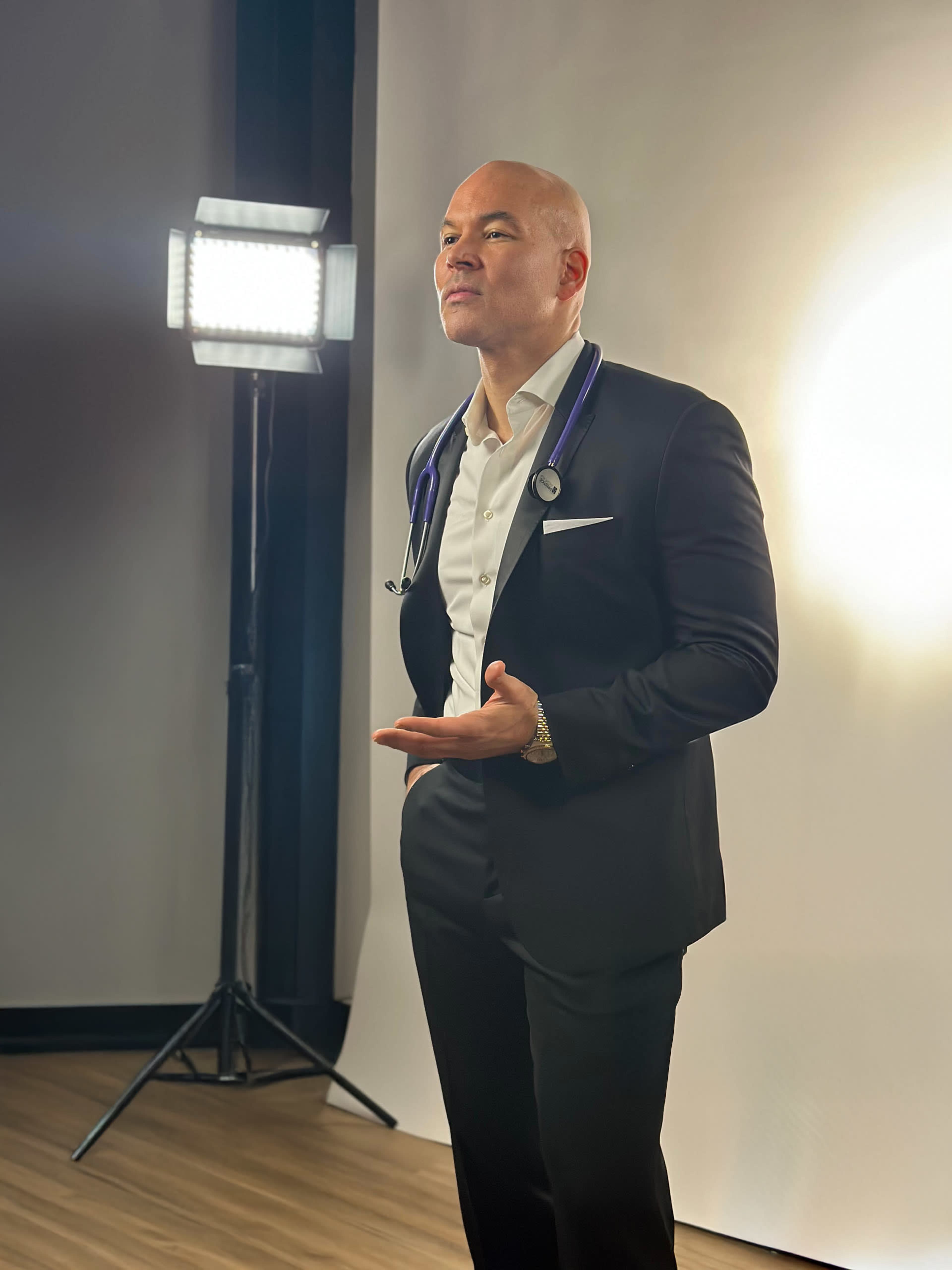 A man in a black suit stands with a stethoscope around his neck in front of a bright studio light.