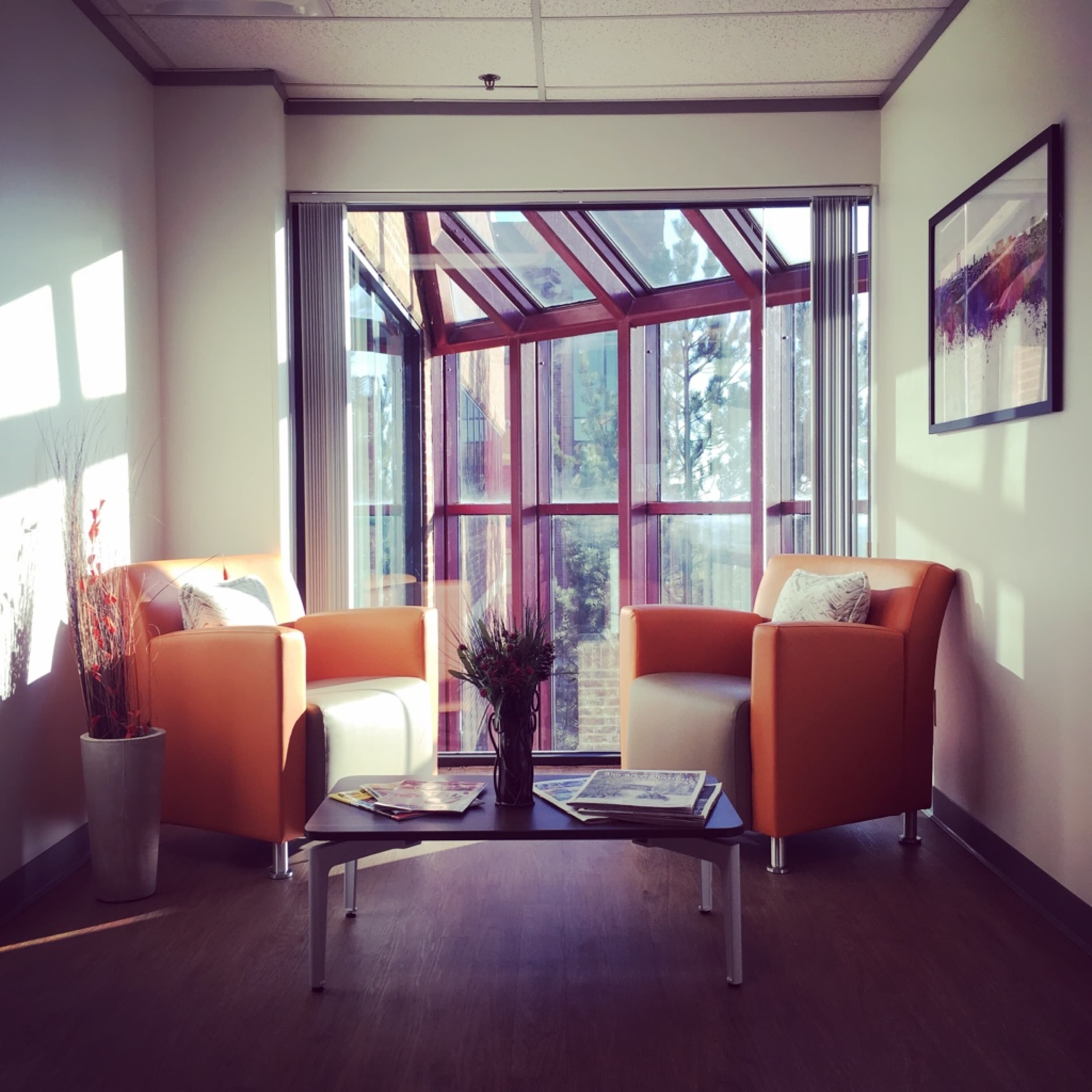 A bright seating area with two orange armchairs facing a glass sectioned wall, featuring a small coffee table with magazines in the center.