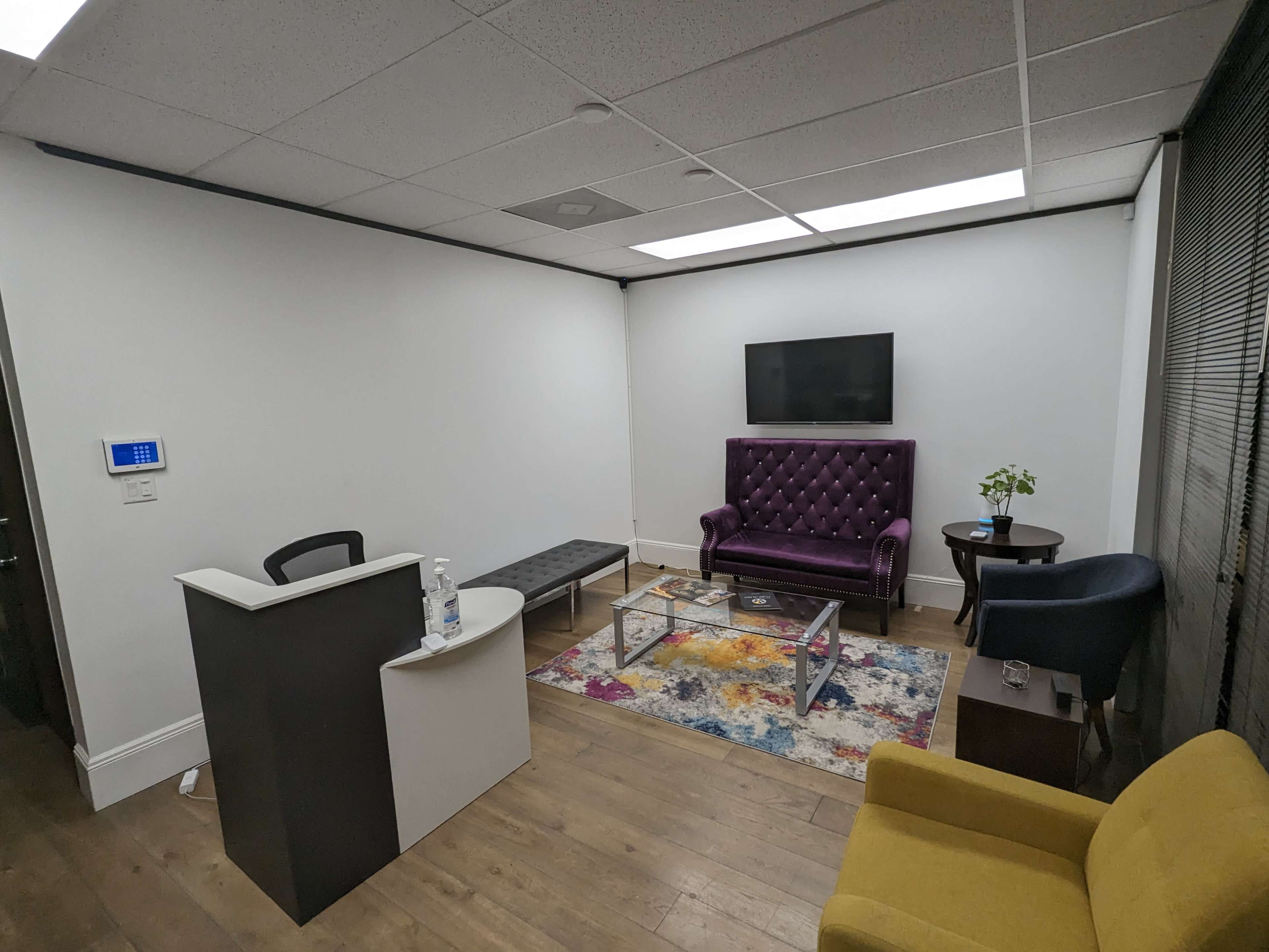 A modern office space with a purple upholstered sofa, a glass coffee table, a small plant on a side table, and a reception area featuring a minimalist desk.