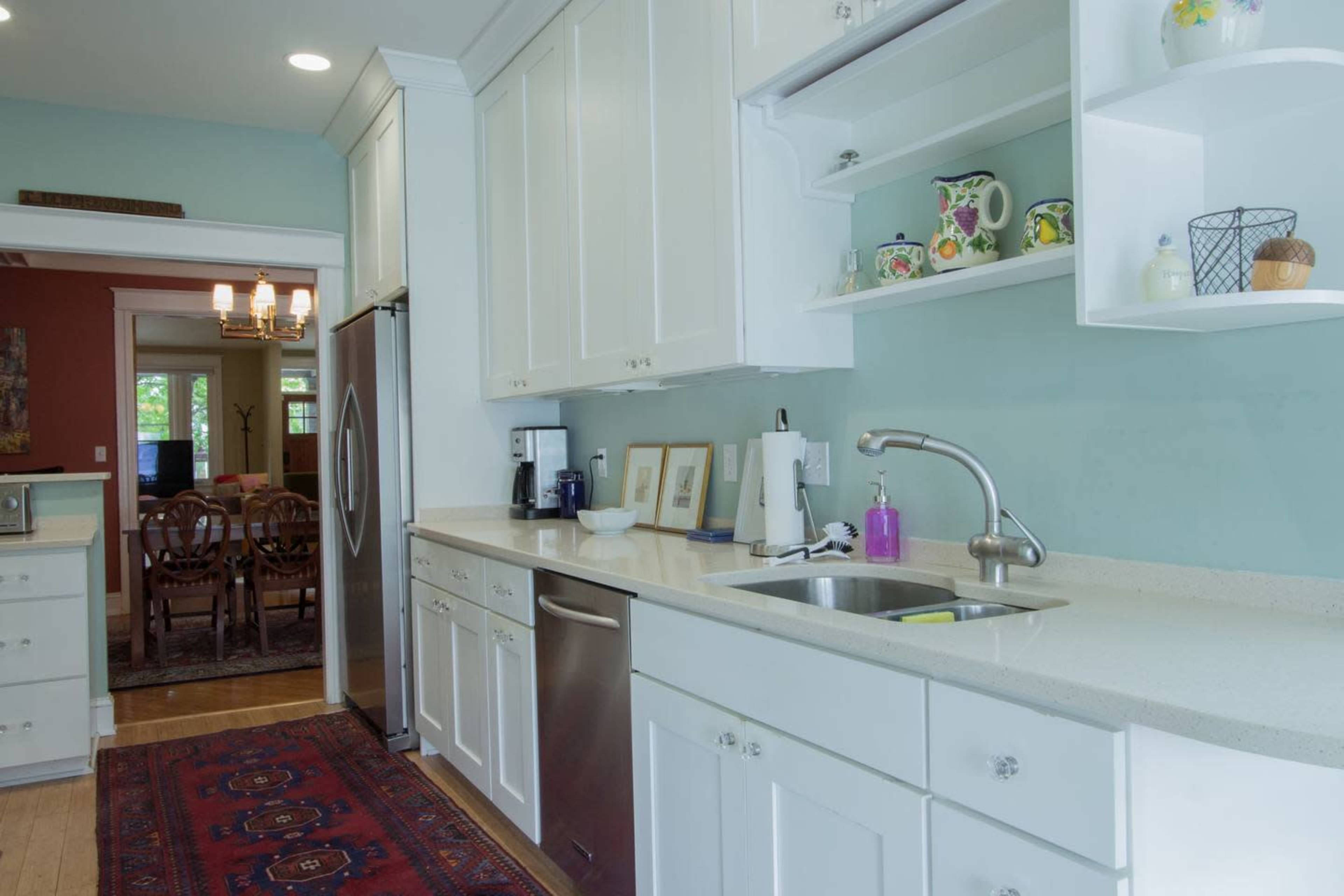 A bright kitchen featuring white cabinetry, a stainless steel refrigerator, and a sink with a countertop, leading to a dining area in the background.