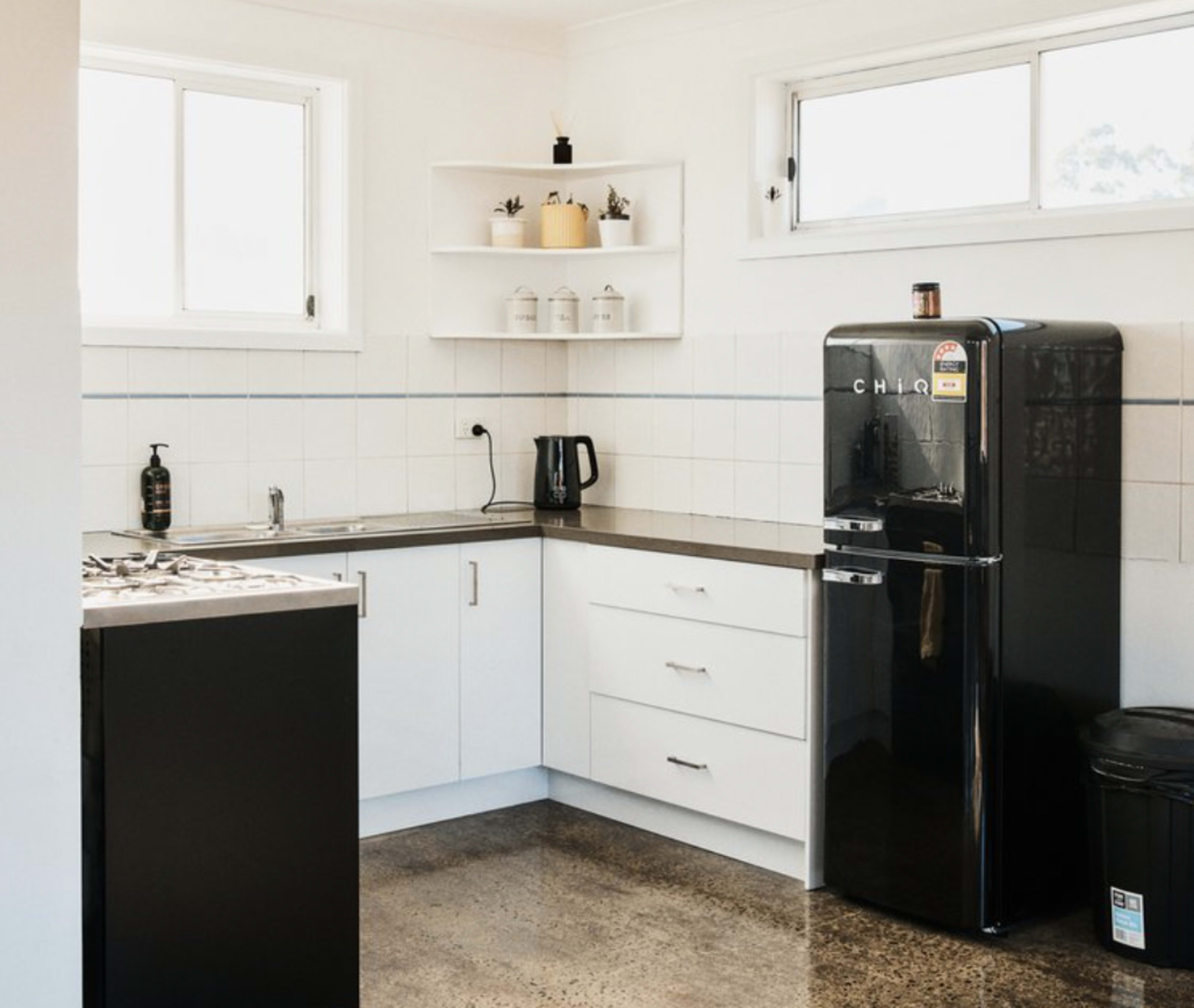 A modern kitchen featuring a black refrigerator, a stainless steel sink, a kettle on the countertop, and white cabinetry against a tiled wall.