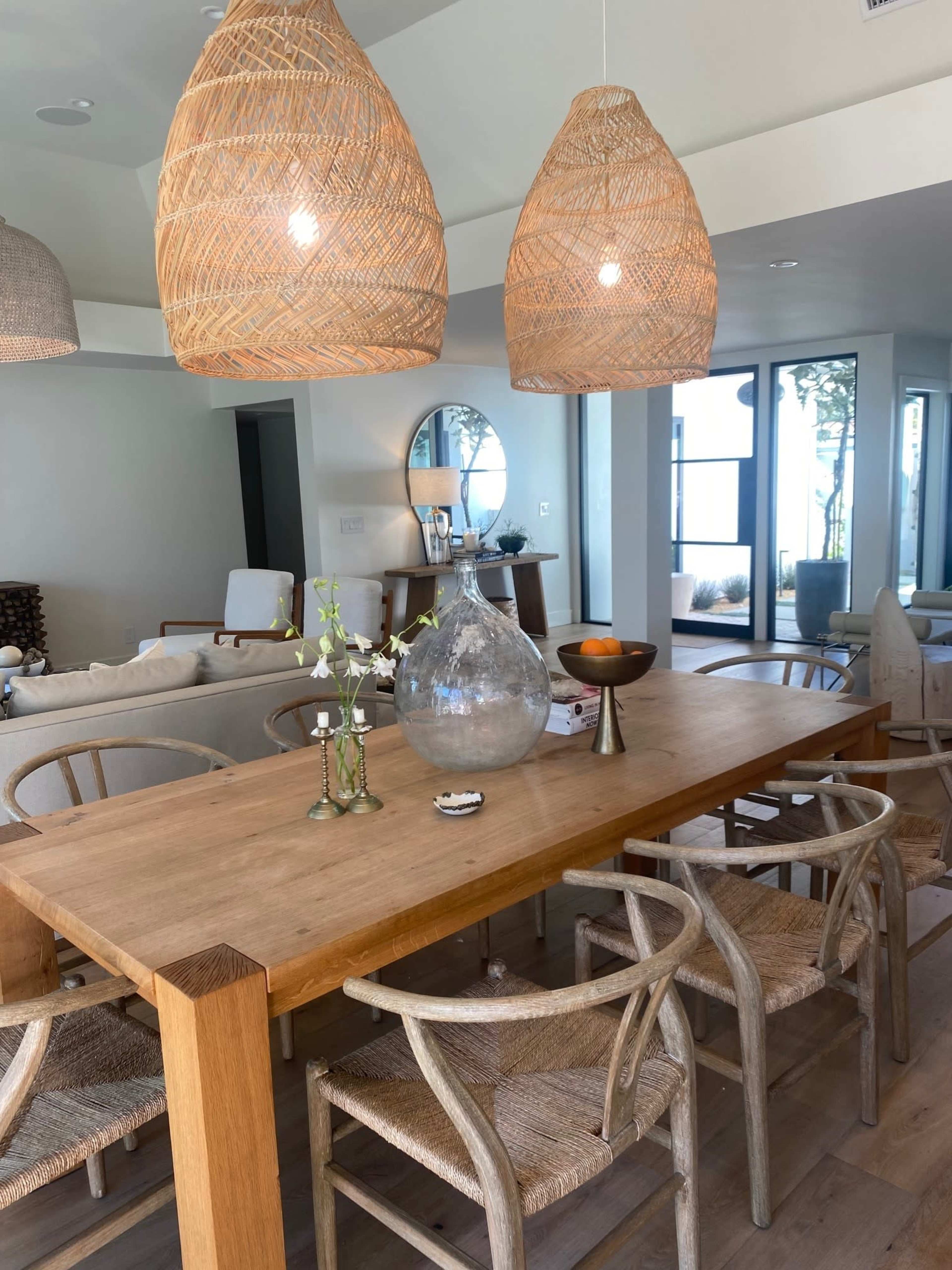 A wooden dining table is set with decorative items and surrounded by light-colored chairs in a modern interior with wicker light fixtures.