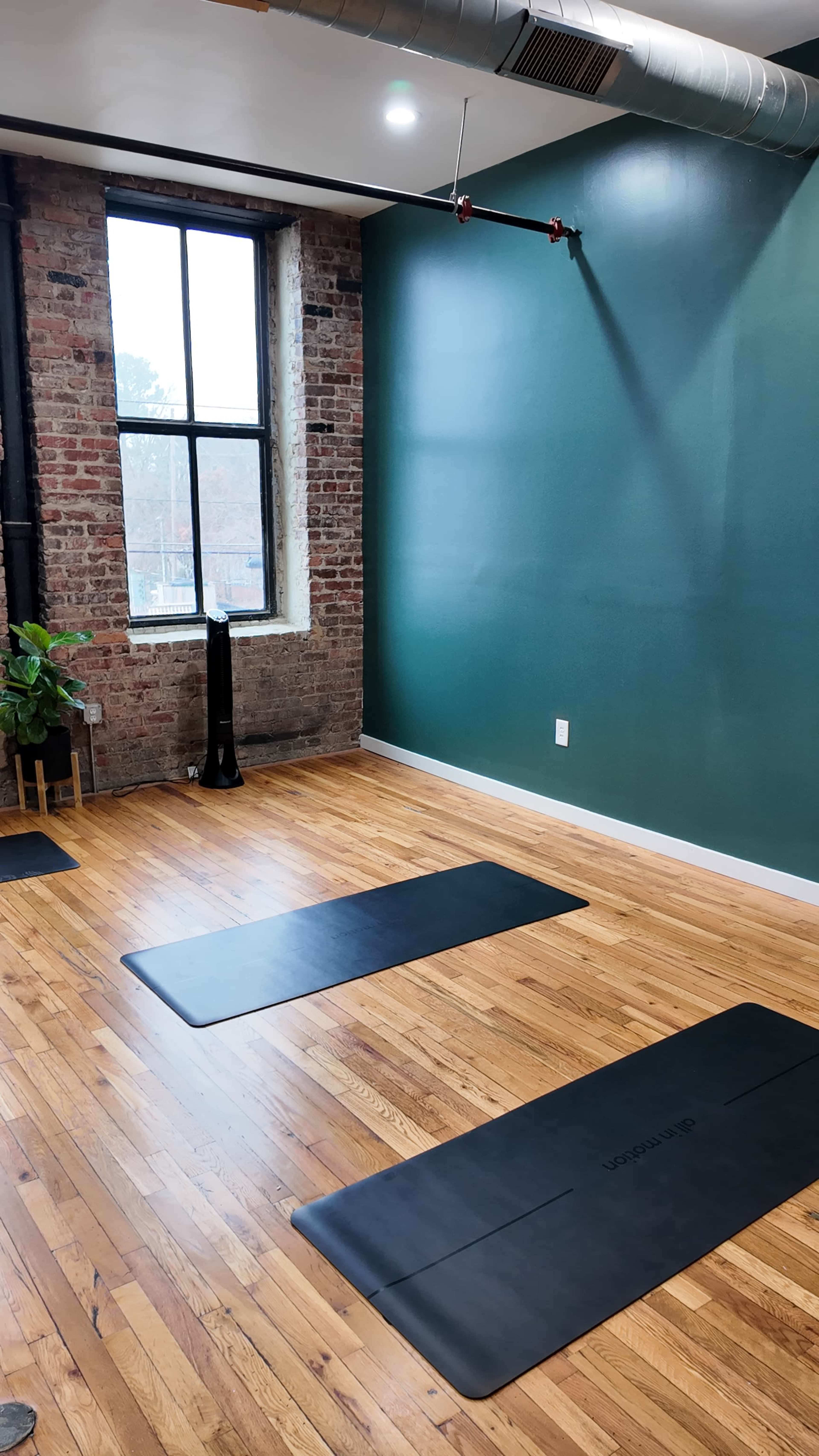 The image shows a yoga studio with hardwood floors, three black mats on the ground, a brick wall, and a large window letting in natural light.