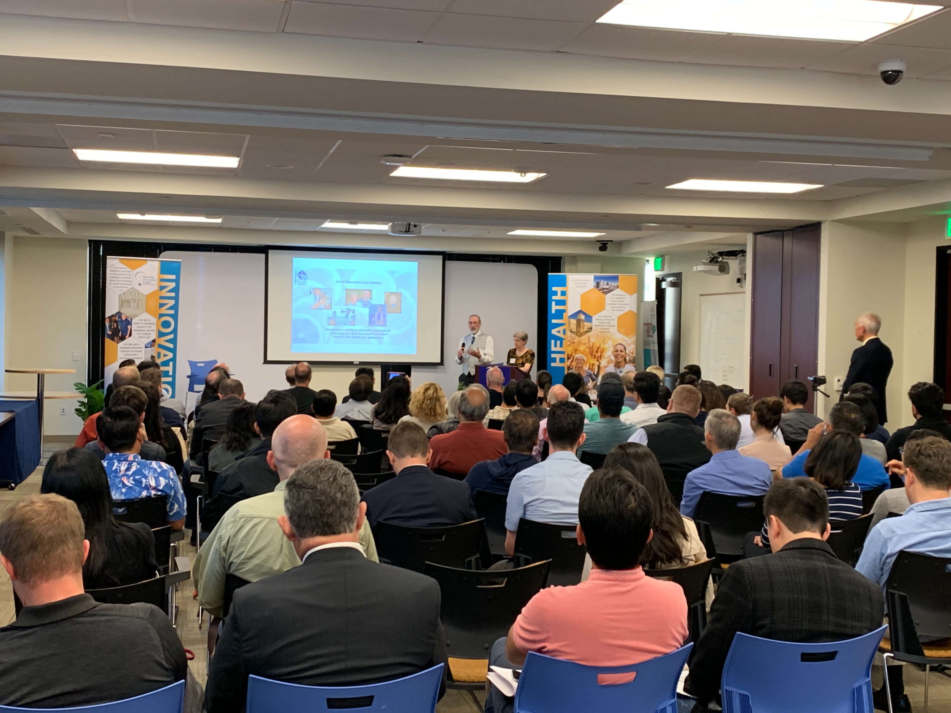 A speaker presents in front of an audience seated in a conference room with banners on either side highlighting innovation in health.