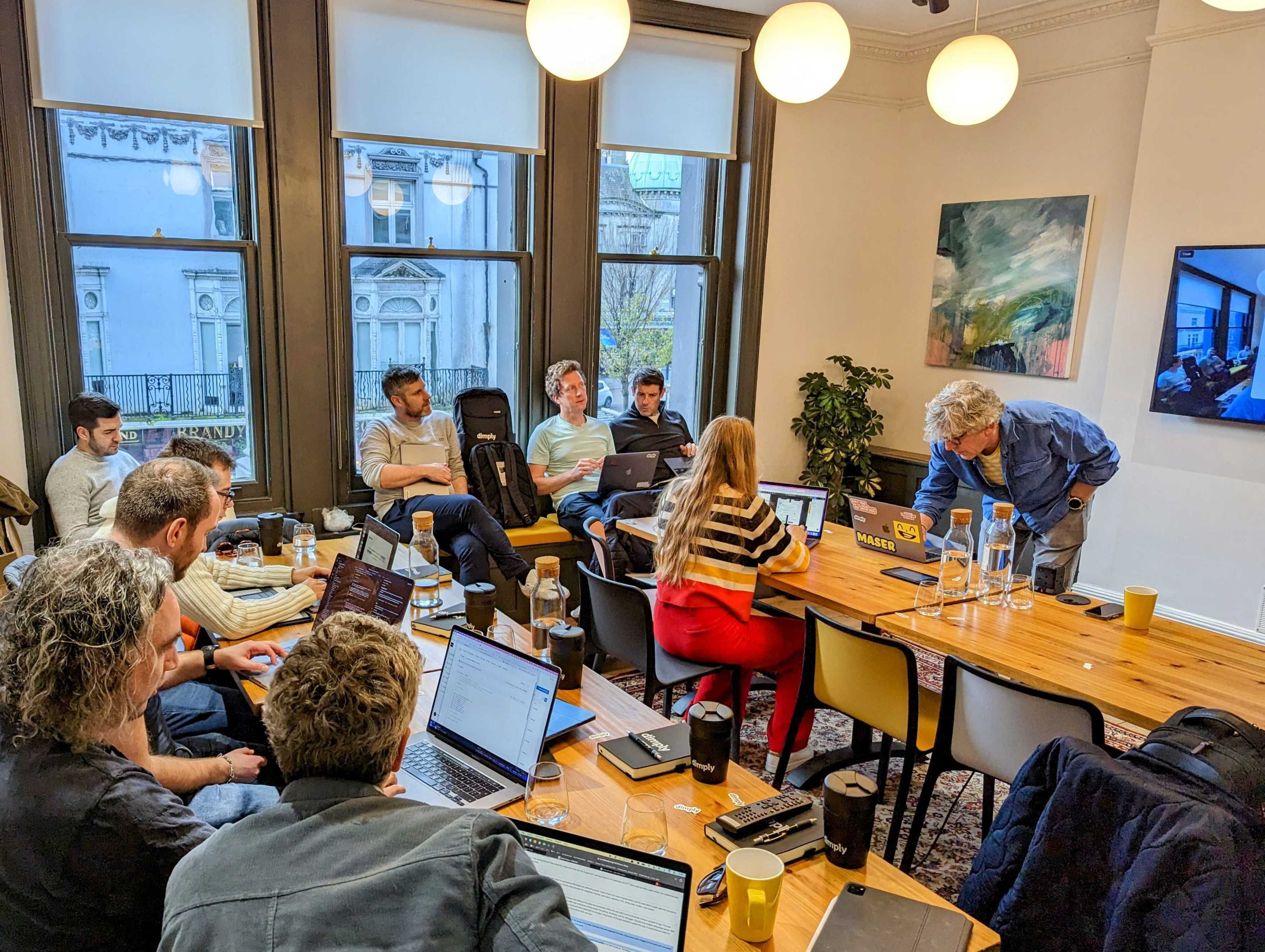A group of people is seated around a wooden table in a meeting room, with laptops open and a presentation displayed on a screen.