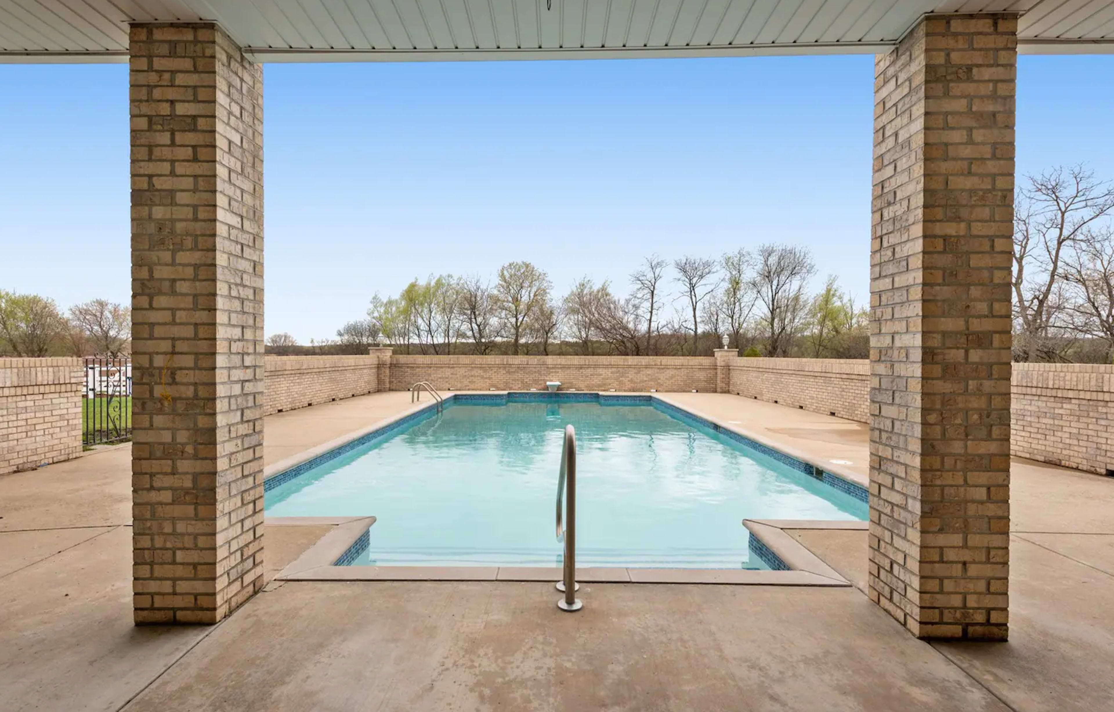 A rectangular swimming pool framed by two brick columns and surrounded by a concrete deck, with trees visible in the background.