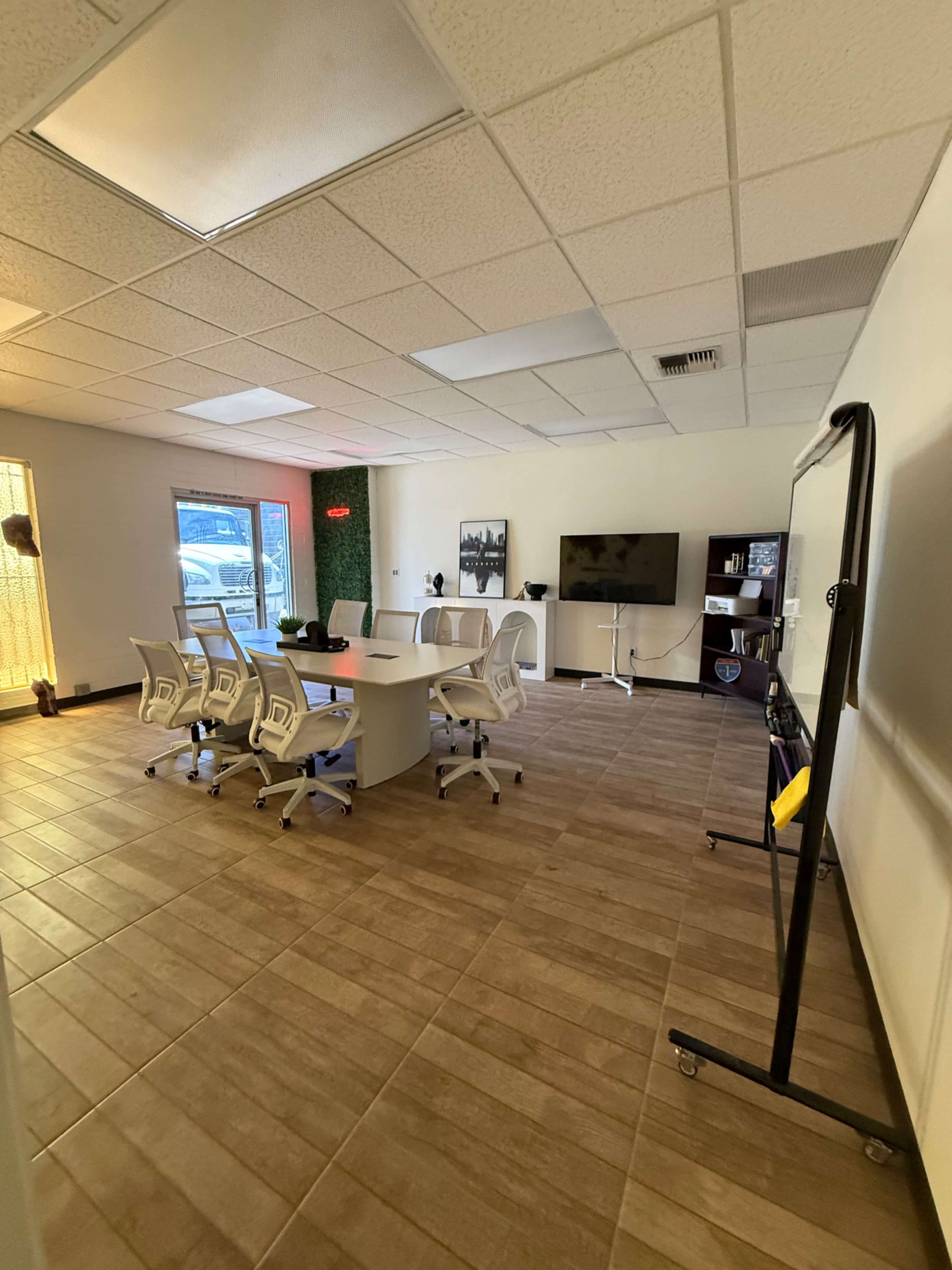 The image shows a conference room with a large round table surrounded by several white office chairs, a television mounted on the wall, and a green accent wall.
