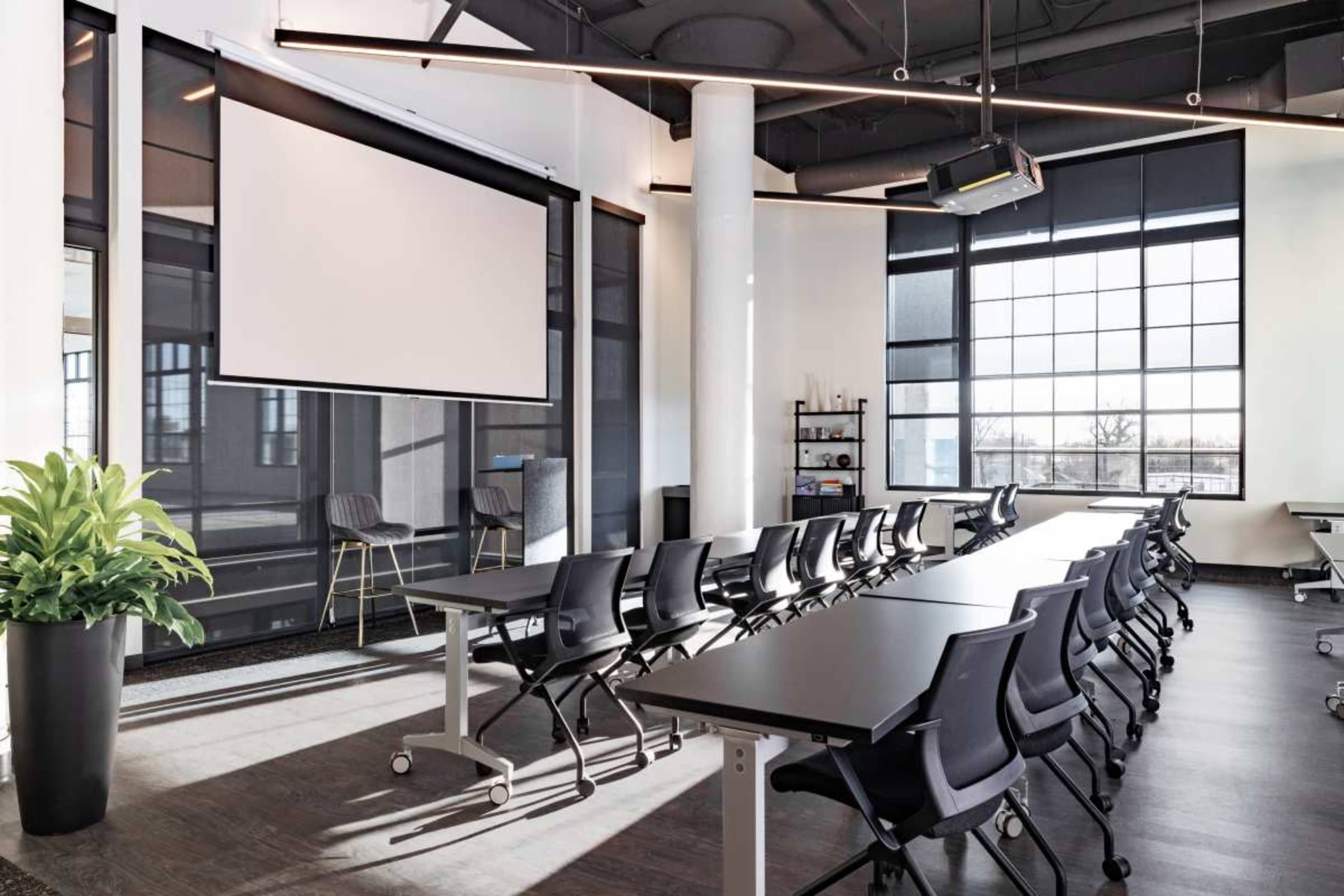The image shows a modern conference room with rows of tables and chairs facing a large projection screen, illuminated by natural light from large windows.