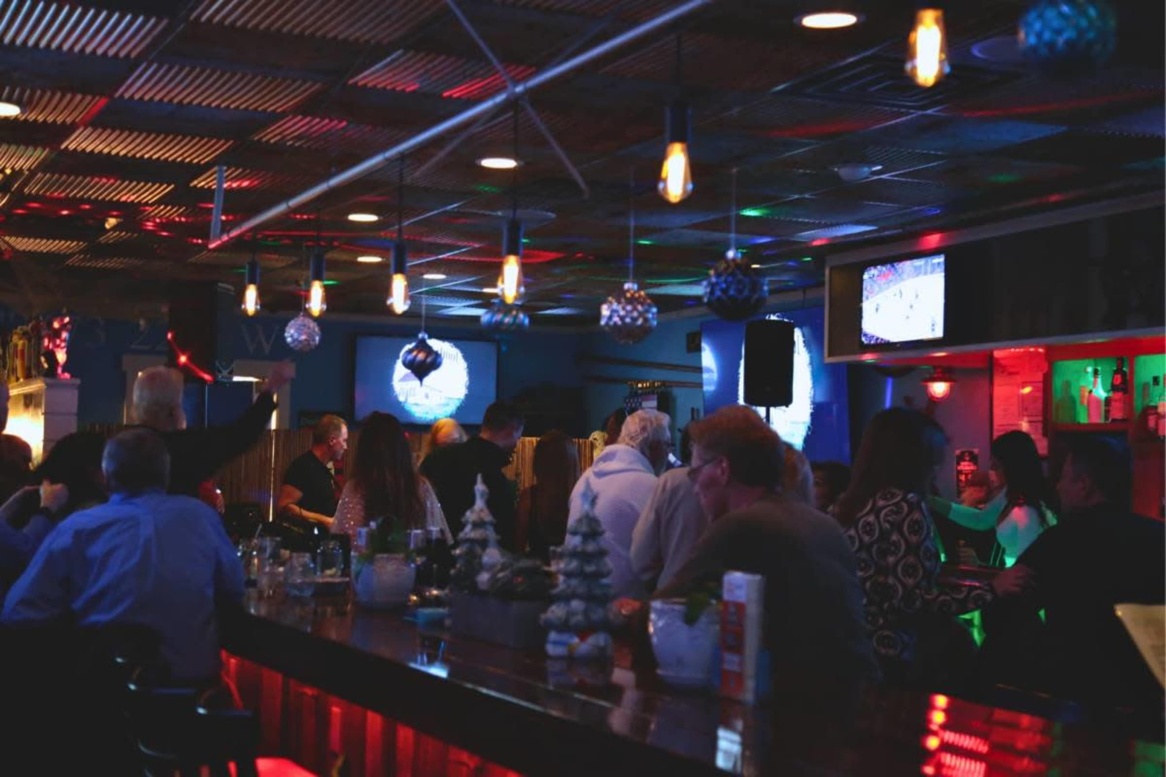 The scene shows a lively bar with patrons socializing under colorful lighting, decorated for the holiday season.