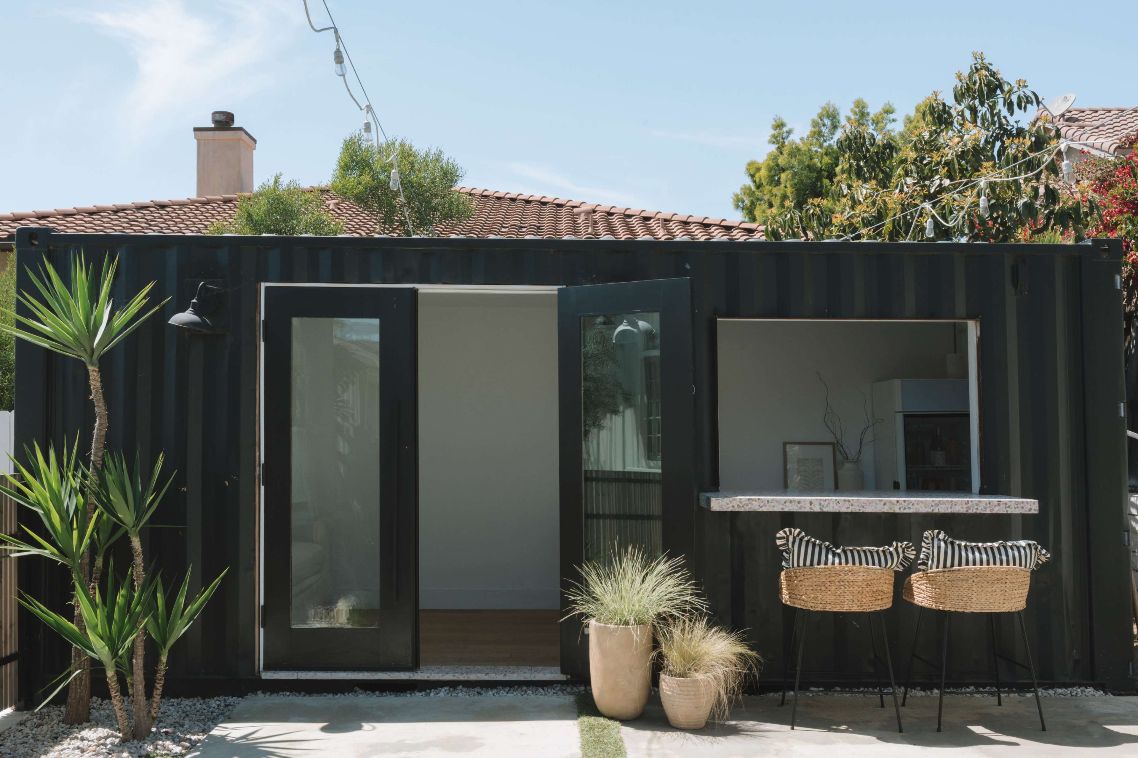 A modern black shipping container serves as a backyard living space, featuring open doors and a small outdoor bar with two chairs surrounded by landscaping.