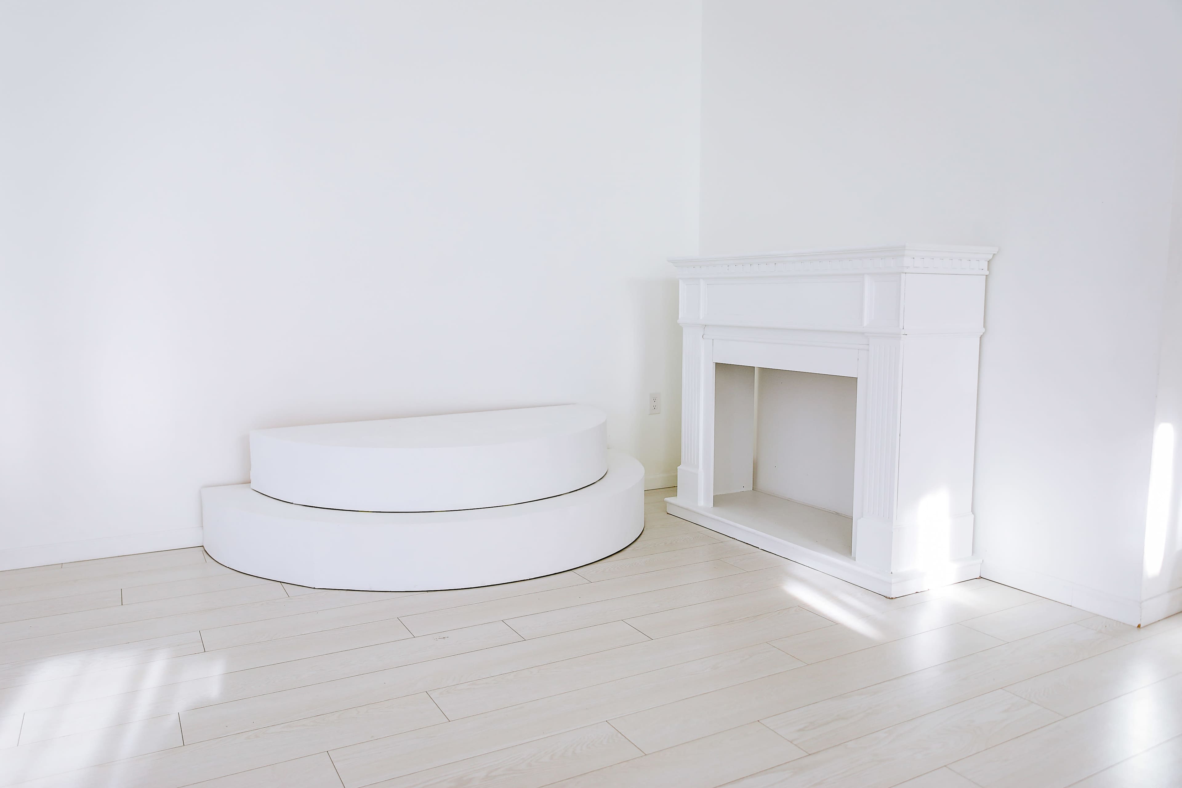 The image shows a minimalist room with a white curved bench and a white fireplace against a bright wall.