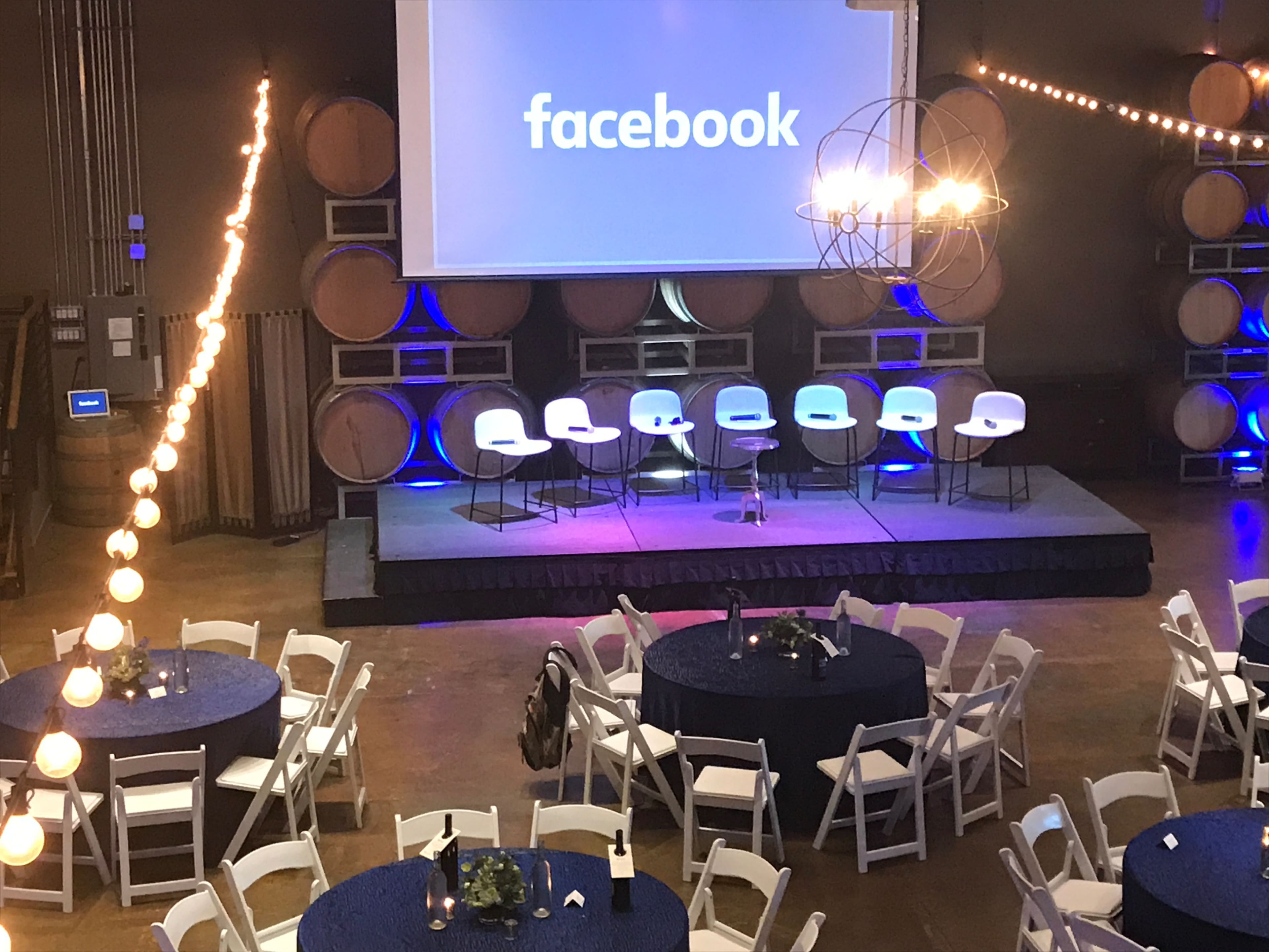 A stage setup for an event in a venue with large wine barrels, featuring a "facebook" logo on a screen and several white chairs arranged behind a table.