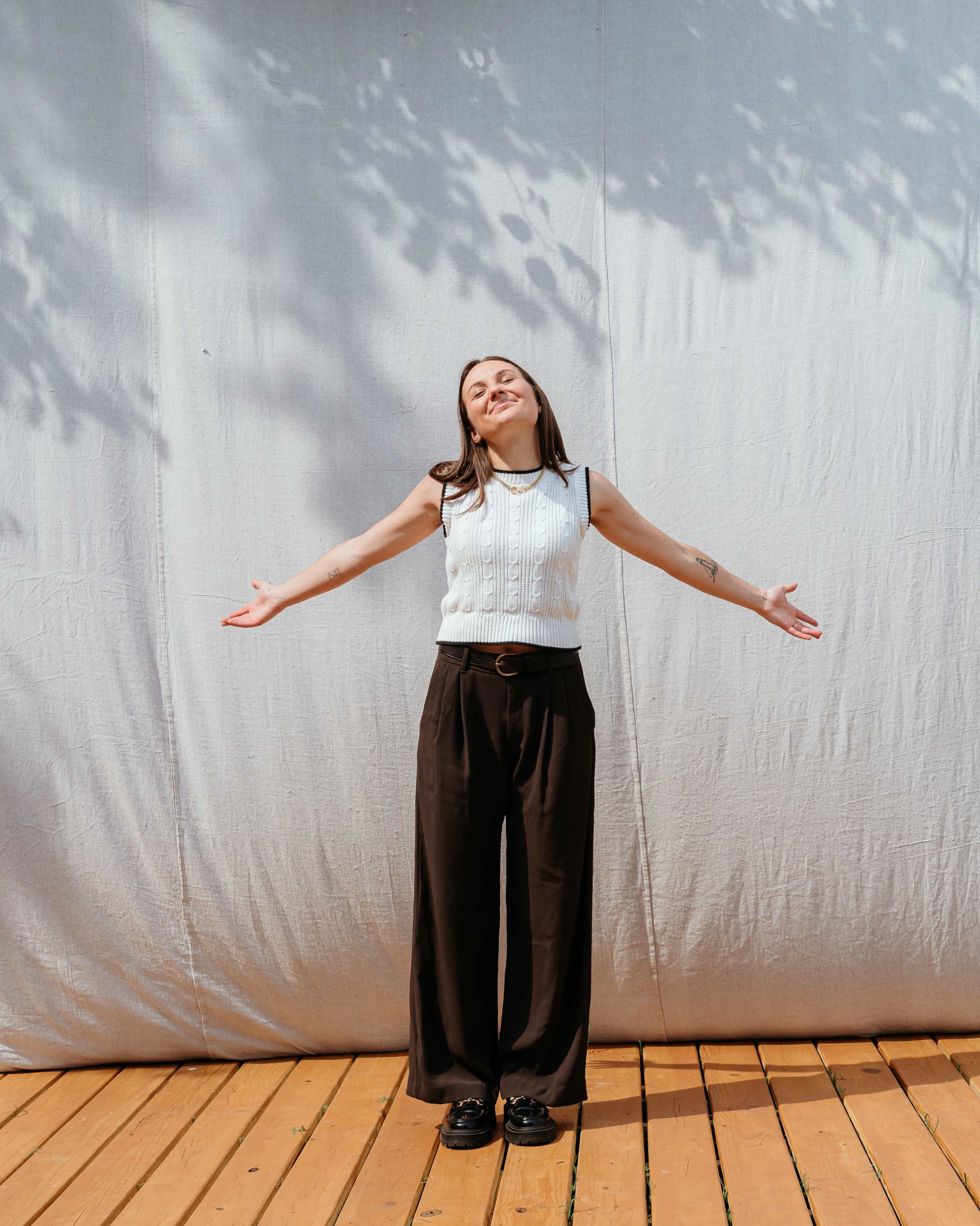 A woman in a sleeveless white sweater and black trousers stands with her arms outstretched in front of a light grey backdrop.