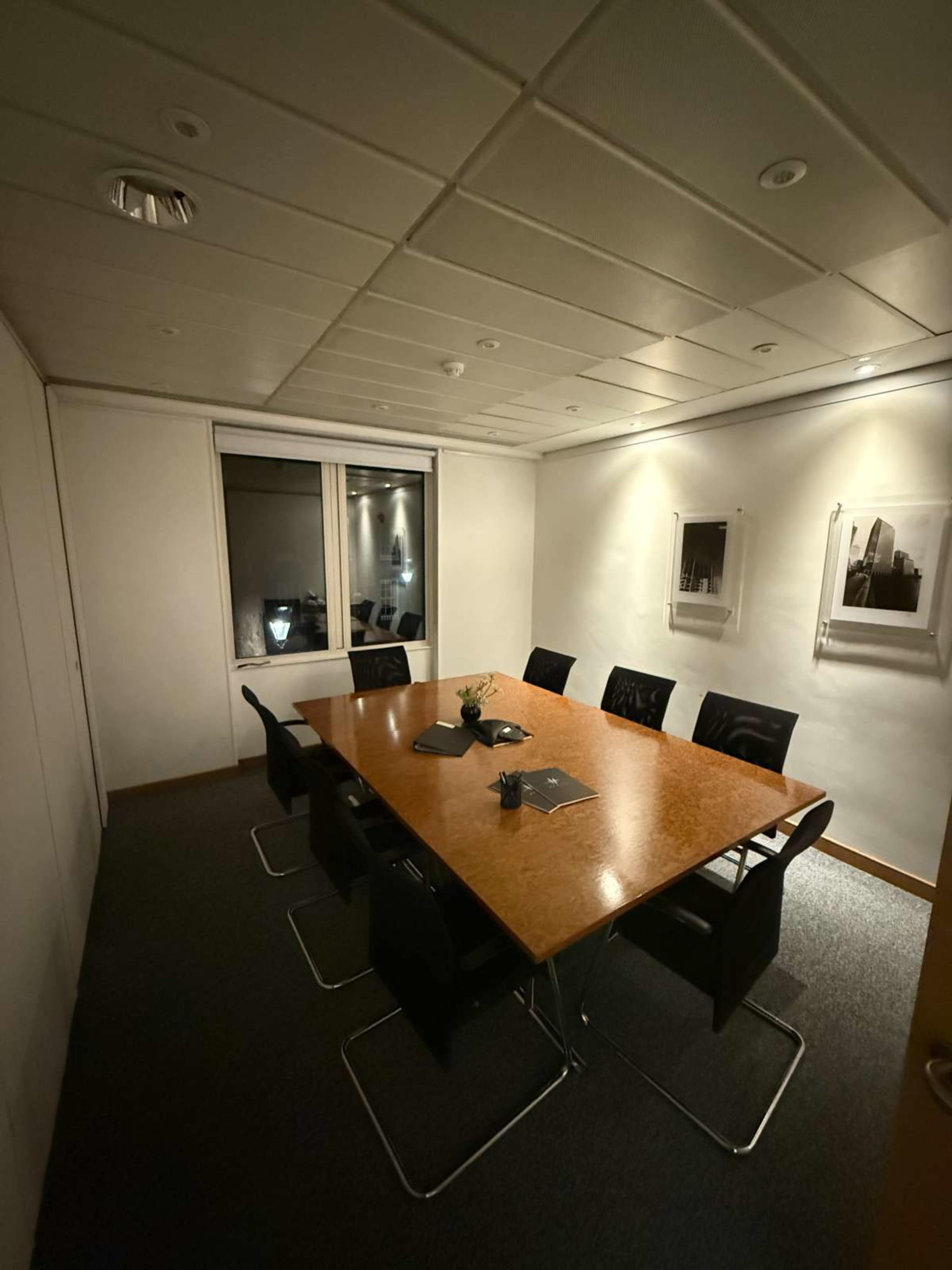 A conference room with a large wooden table surrounded by black chairs, featuring a window and framed photographs on the walls.